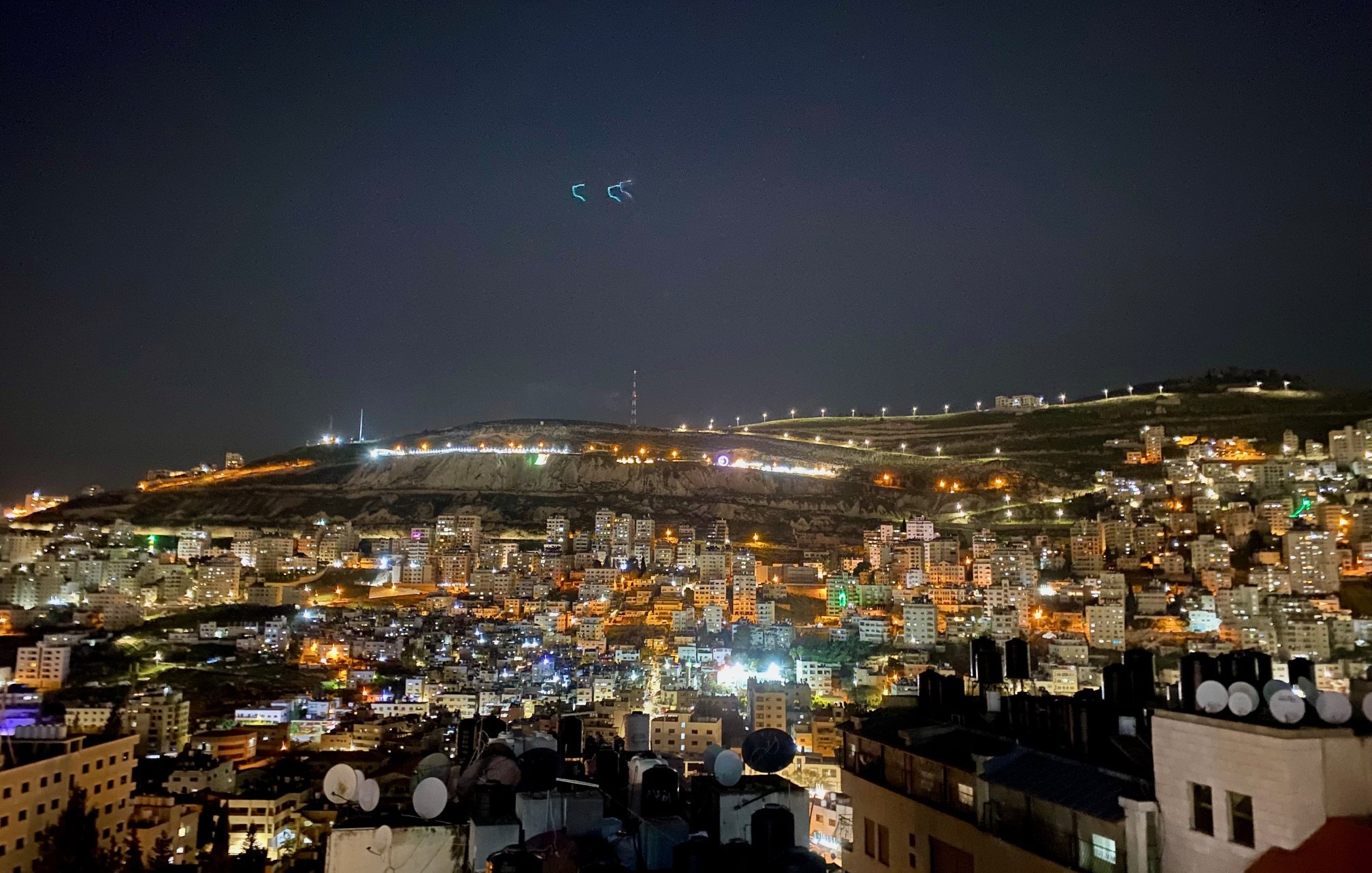 L'image montre une vue nocturne d'une ville illuminée. On peut voir de nombreux bâtiments agencés en terrasses, avec des lumières qui scintillent dans l'obscurité. À l'arrière-plan, des collines sont éclairées, et quelques structures sont visibles au sommet. L'atmosphère est tranquille, avec un ciel nocturne clair. Les antennes et structures en premier plan ajoutent une dimension urbaine à cette scène paisible.