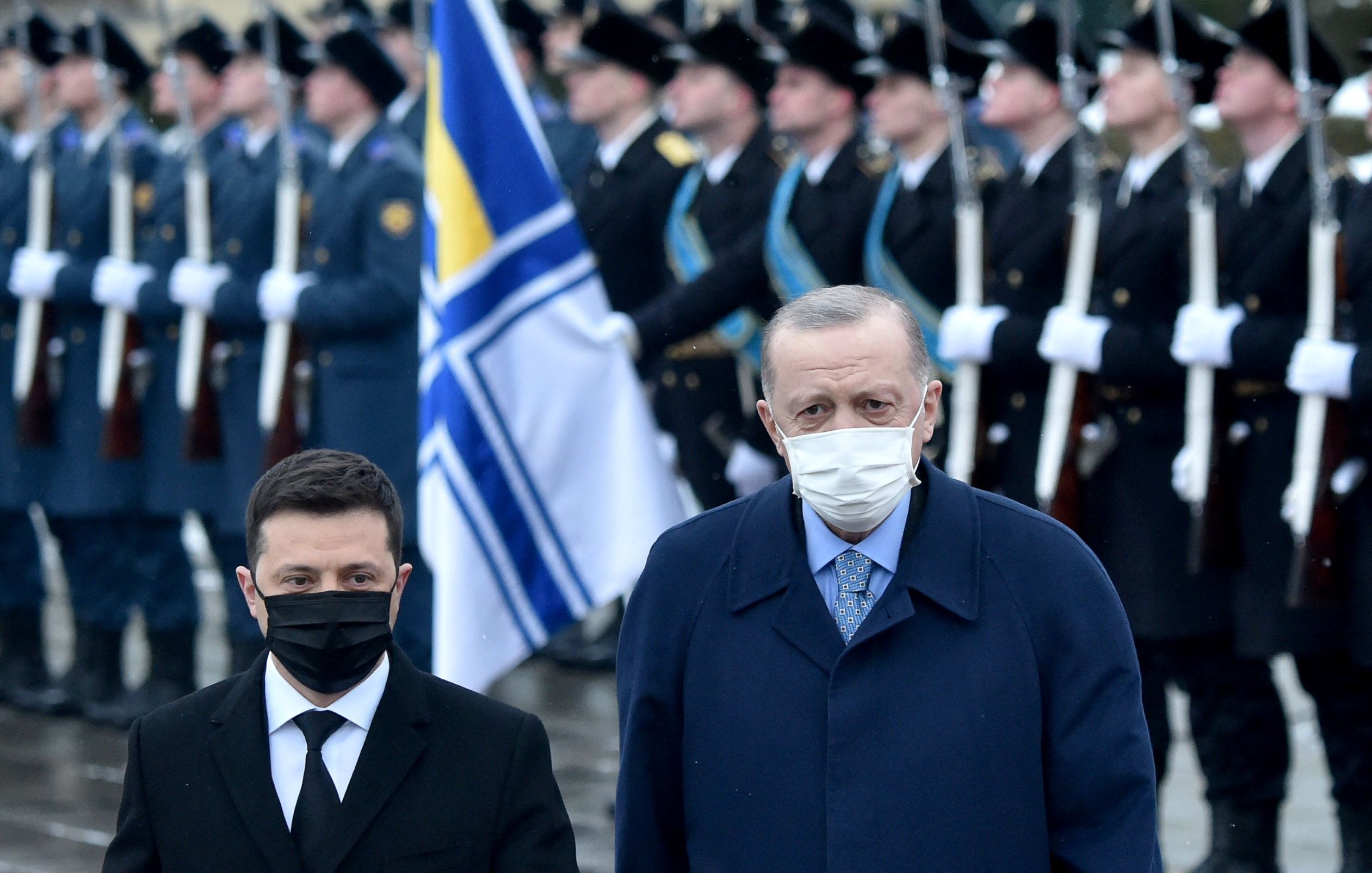 L'image montre deux hommes au premier plan, portant des masques. L'un d'eux est en costume noir, tandis que l'autre est habillé d'un manteau bleu. Ils se tiennent côte à côte, faisant face à une cérémonie militaire en arrière-plan, où des soldats en uniforme se tiennent au garde-à-vous avec des drapeaux. L'atmosphère semble officielle et solennelle.