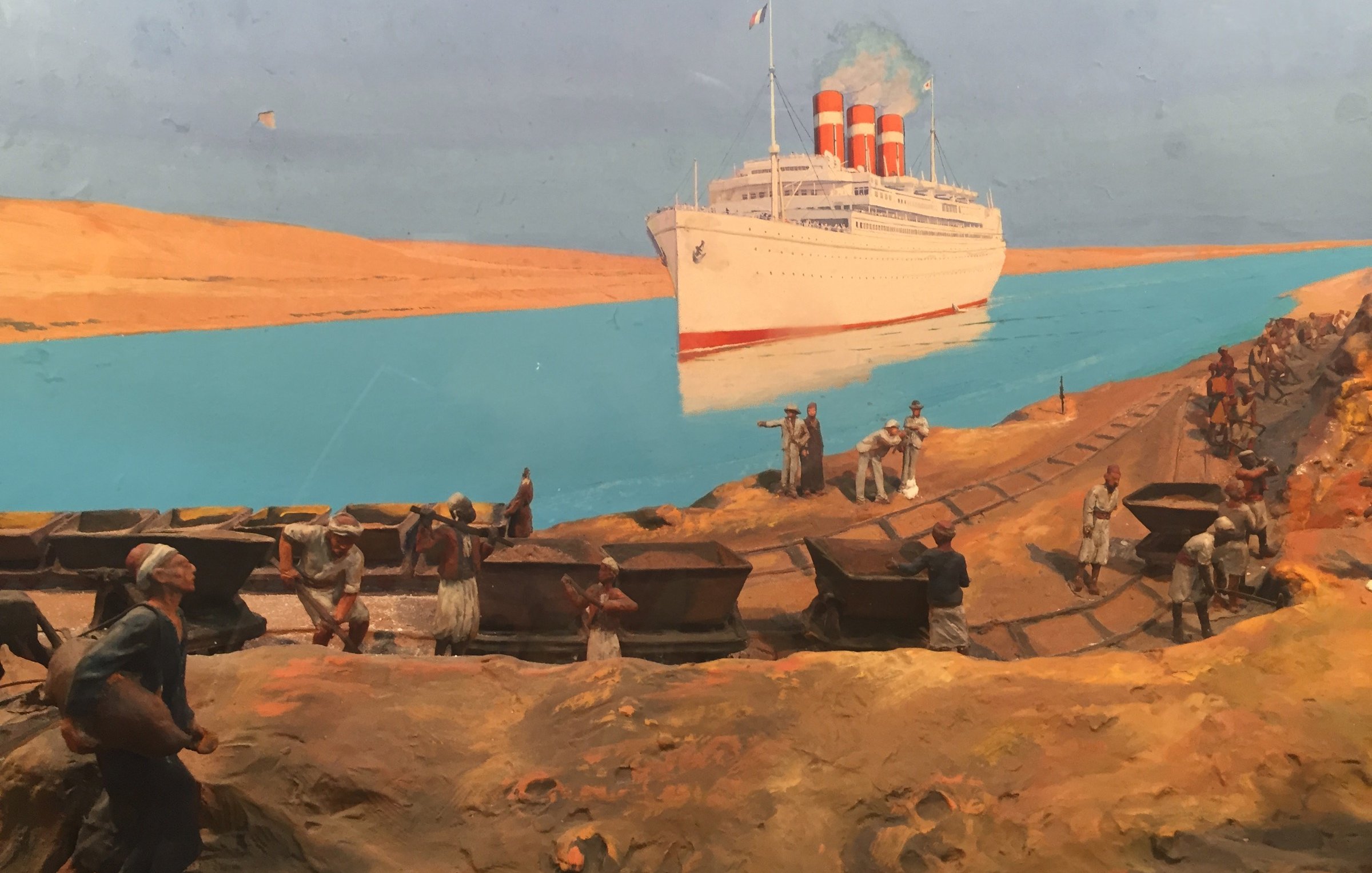 L'image représente une scène historique dans un environnement maritime. On voit un grand paquebot avec trois cheminées rouges amarré près d'une berge, probablement dans un canal ou un fleuve. Au premier plan, plusieurs hommes travaillent sur la rive, transportant des charges, probablement des matériaux ou des marchandises. À l'arrière-plan, le paysage désertique montre des dunes de sable sous un ciel bleu, créant un contraste avec l'eau turquoise. Cette image évoque l'activité maritime et le travail acharné des ouvriers dans un contexte industriel.