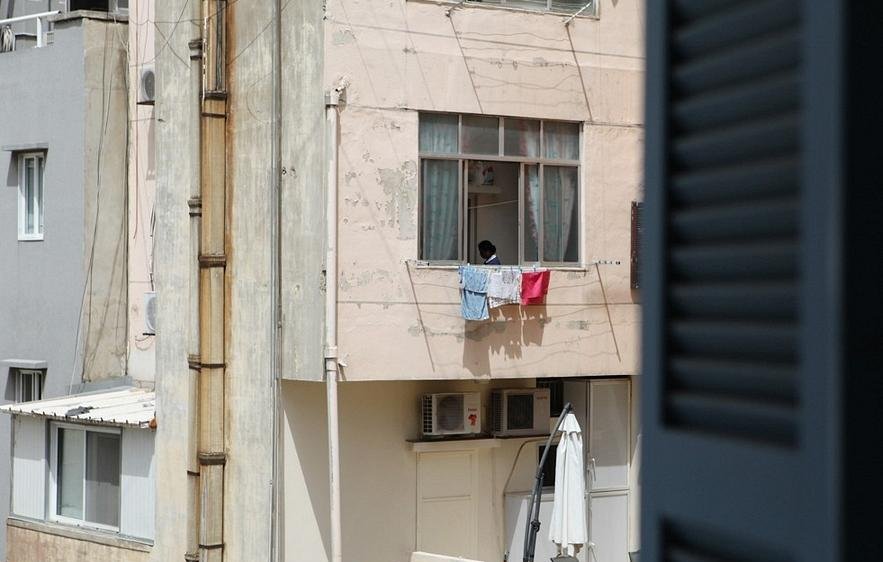 L'image montre un immeuble avec des murs de couleur rose. À un des balcons, on aperçoit un enfant qui semble regarder à l'extérieur. Des vêtements sont étendus sur la rambarde du balcon, et au sol, on peut voir un parasol ouvert. À droite, un volet est partiellement ouvert, ajoutant une autre dimension à la scène. L'ambiance semble calme et typique d'un environnement urbain.