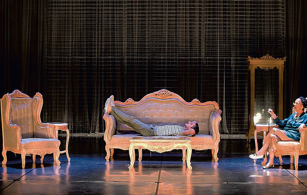 L'image montre une scène de théâtre avec un décor composé de plusieurs meubles anciens. Au centre, un sofa sur lequel un homme est allongé, semble détendu. À droite, une femme est assise sur une chaise, elle semble engagée dans une conversation ou une réflexion. Le fond est constitué d'un rideau sombre, ce qui met en valeur les meubles éclairés. L'ambiance semble à la fois intime et dramatique, suggérant une interaction entre les deux personnages.