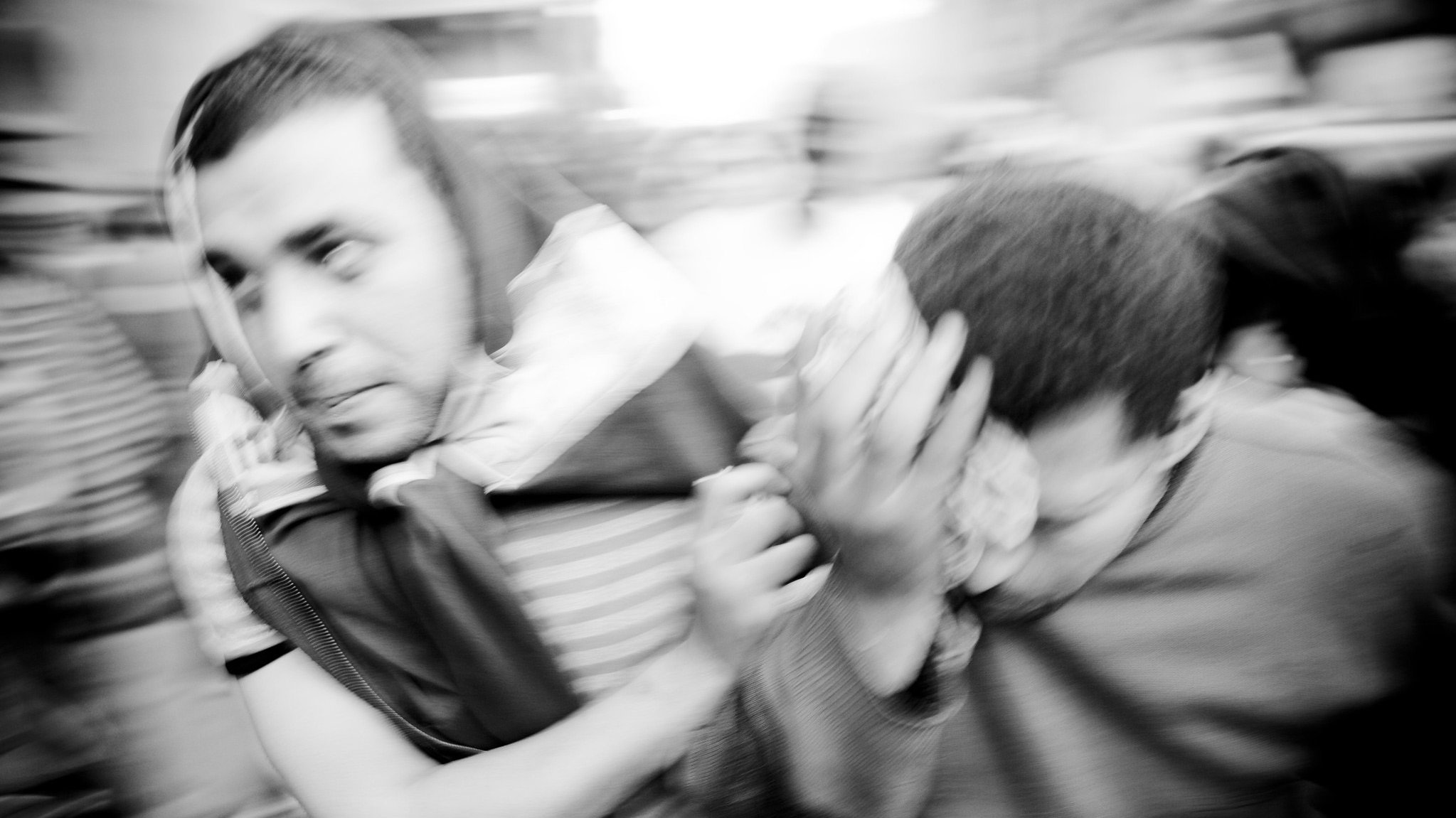 L'image montre deux personnes en mouvement dans un environnement flou, ce qui donne une impression de dynamisme et d'urgence. L'une des personnes semble porter quelque chose sur son épaule, tandis que l'autre se couvre le visage avec une main. L'atmosphère est intense, comme si les sujets étaient en train de fuir ou de réagir à une situation pressante. Les couleurs sont principalement en noir et blanc, accentuant le contraste et l'émotion de la scène.