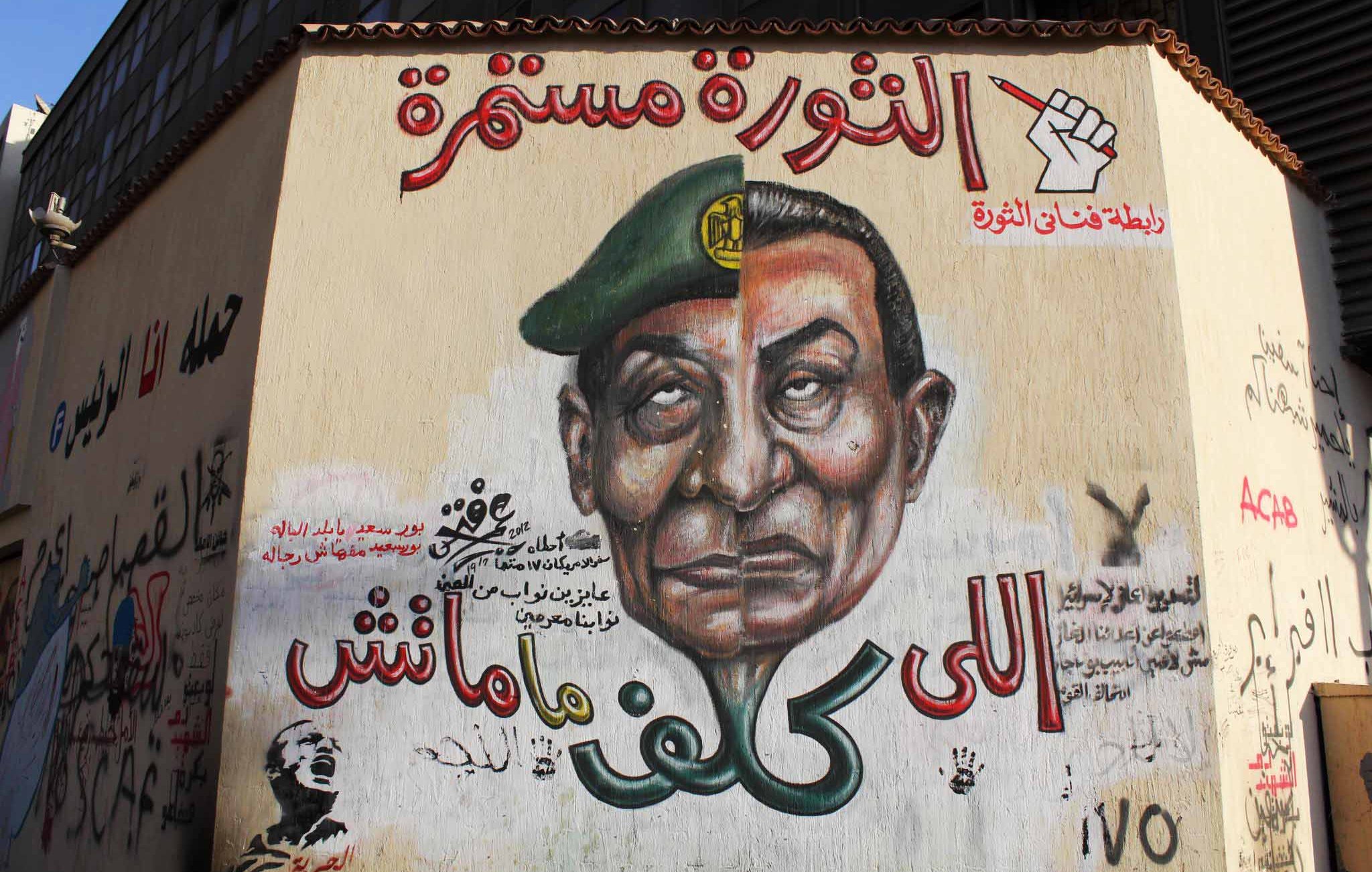 L'image montre une fresque murale sur un mur, représentant un homme avec un uniforme militaire et un képi. Son expression est sérieuse et emblématique. Autour de cette image, on trouve des inscriptions en arabe, dont certaines font allusion à une continuité de la révolution. Le style artistique est vibrant et engagé, évoquant des thèmes de soulèvement et de résistance. Des symboles de révolte ou de mouvement peuvent également être présents, soulignant un message politique fort.