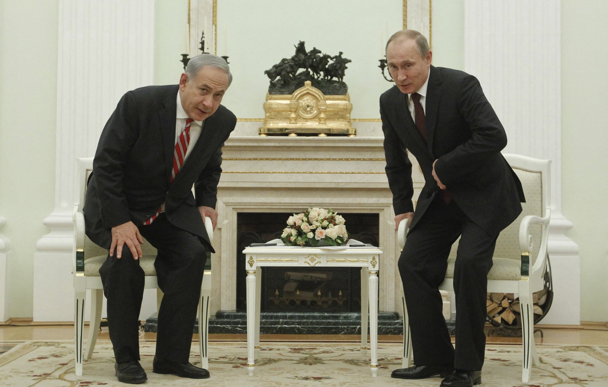 L'image montre deux hommes en costume, l'un avec une cravate rayée et l'autre avec une cravate sombre, se tenant debout devant une table basse dans une pièce élégante. Ils semblent se pencher légèrement en avant, probablement lors d'une conversation. En arrière-plan, on peut voir des éléments de décoration intérieure, dont une grande horloge et un bouquet de fleurs sur la table. Les murs sont peints dans des tons clairs et l'ensemble donne une impression de formalité.