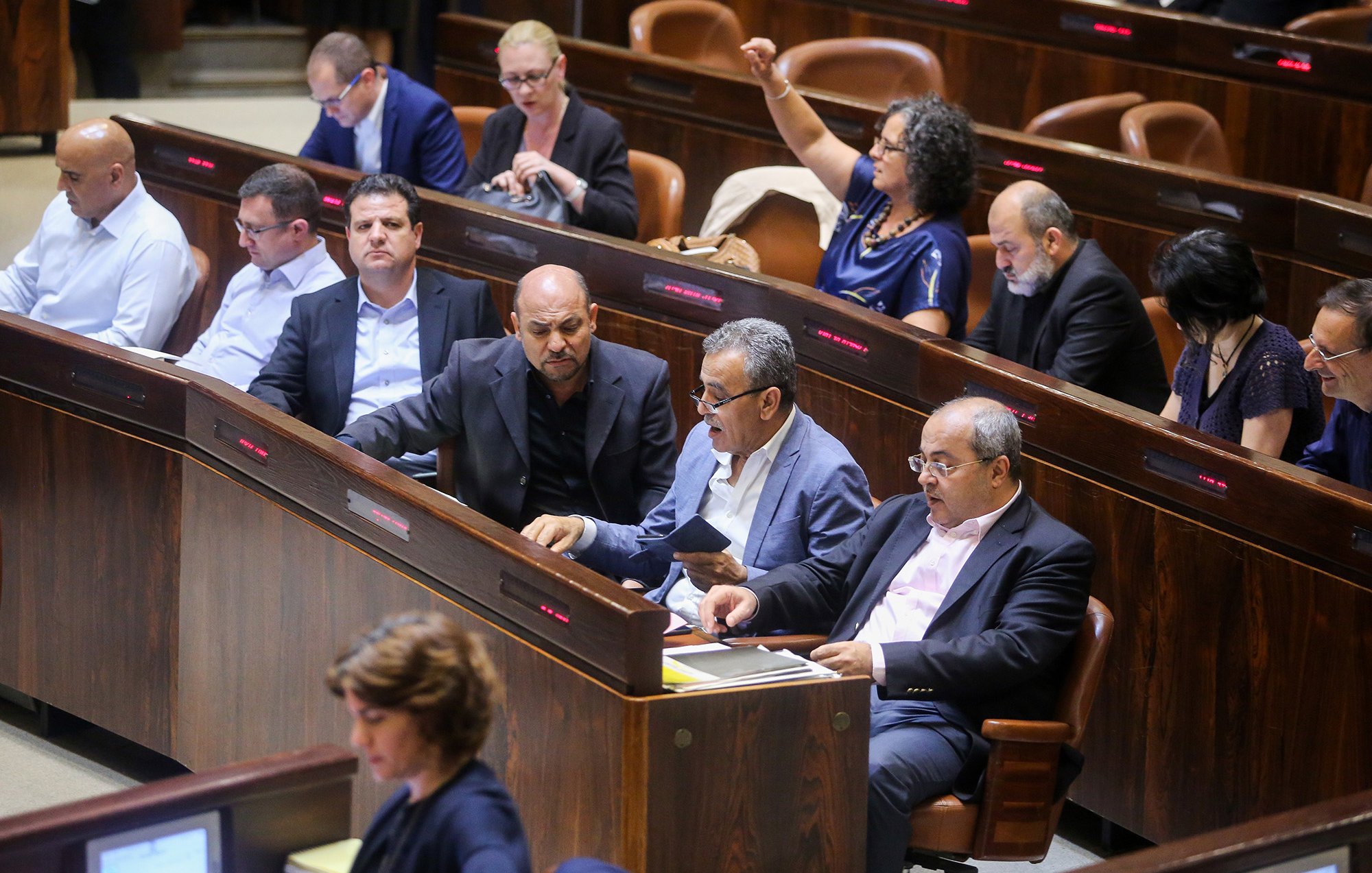 L'image montre une scène à l'intérieur d'une assemblée, probablement dans un parlement. On peut voir plusieurs personnes assises dans des rangées, en train de discuter ou de prendre des notes. Certaines semblent attentives, tandis que d'autres sont engagées dans des conversations. L'environnement est formel, avec des sièges en bois et un éclairage tamisé. Cette composition indique un moment de débat ou de discussion politique.