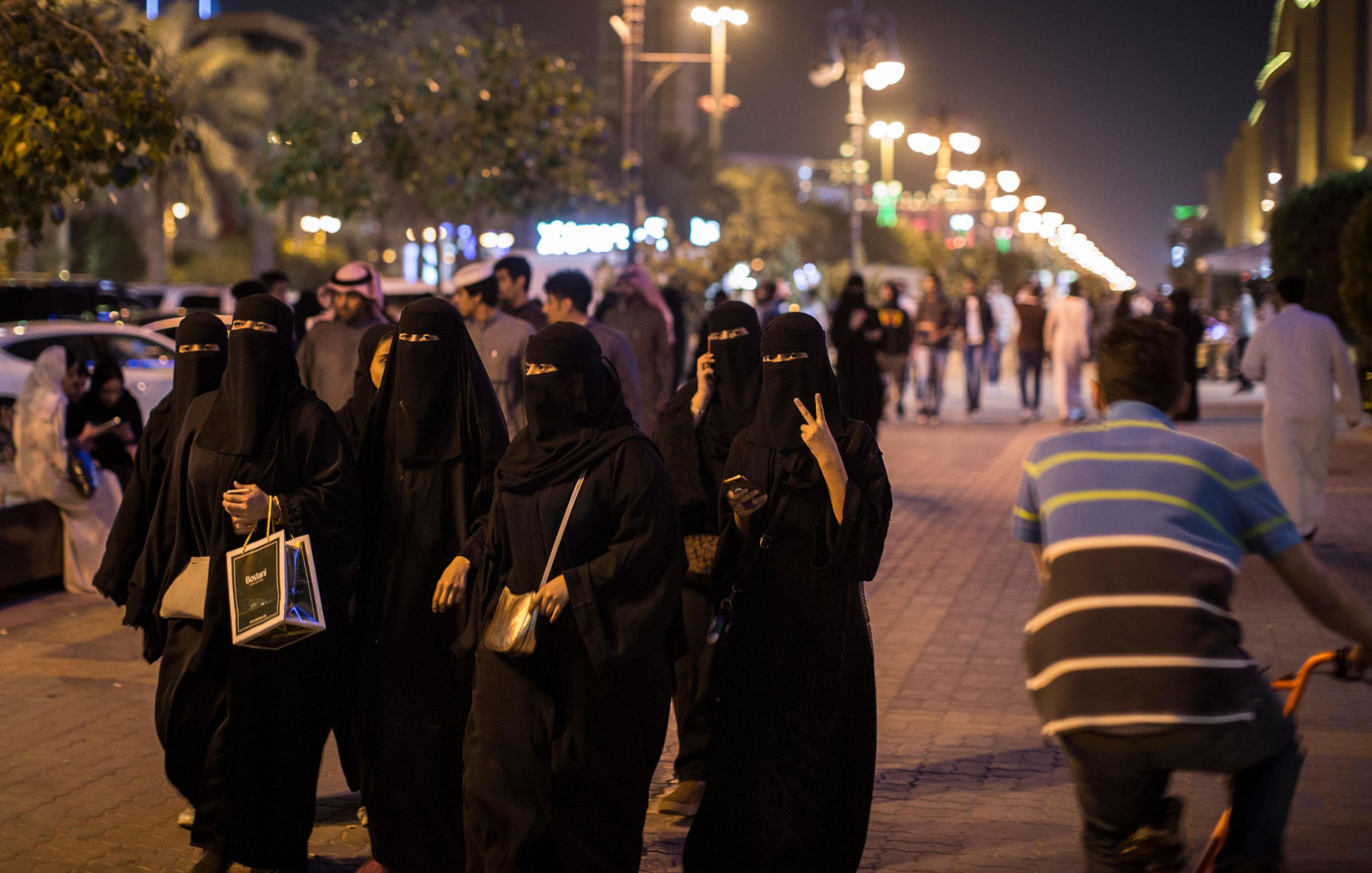 L'image montre un groupe de femmes vêtues de tenues noires traditionnelles marchant ensemble sur un trottoir. Certaines d'entre elles portent des masques qui dissimulent leur visage, tandis que d'autres affichent des gestes amicaux. En arrière-plan, on voit une rue animée avec des passants et des lumières, évoquant une ambiance nocturne. Un homme à vélo passe à proximité, ajoutant à la dynamique de la scène.