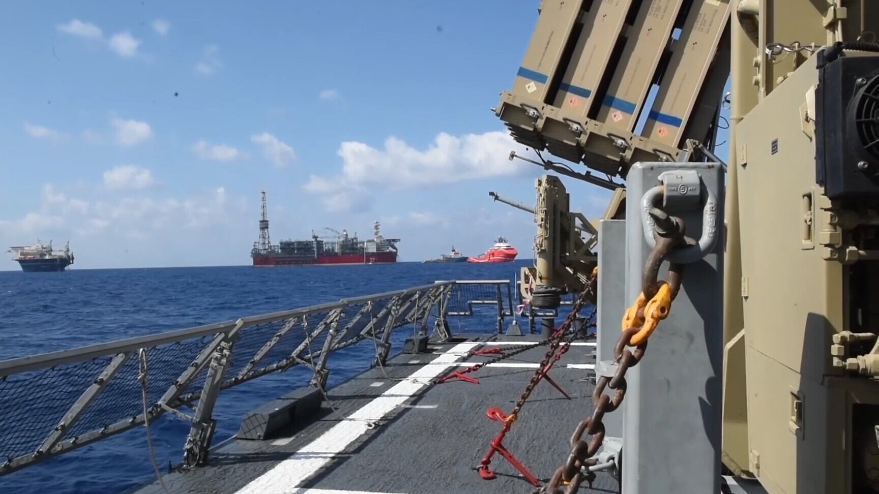 L'image montre un navire en mer, avec des structures de forage pétrolier visibles à l'horizon. On peut apercevoir un système de défense intégrée, probablement dédié à la surveillance ou à la protection, installé sur le pont du navire. Le ciel est clair avec quelques nuages, et la mer est calme, créant une atmosphère maritime paisible. Des chaînes et des équipements de sécurité sont également visibles sur le pont.