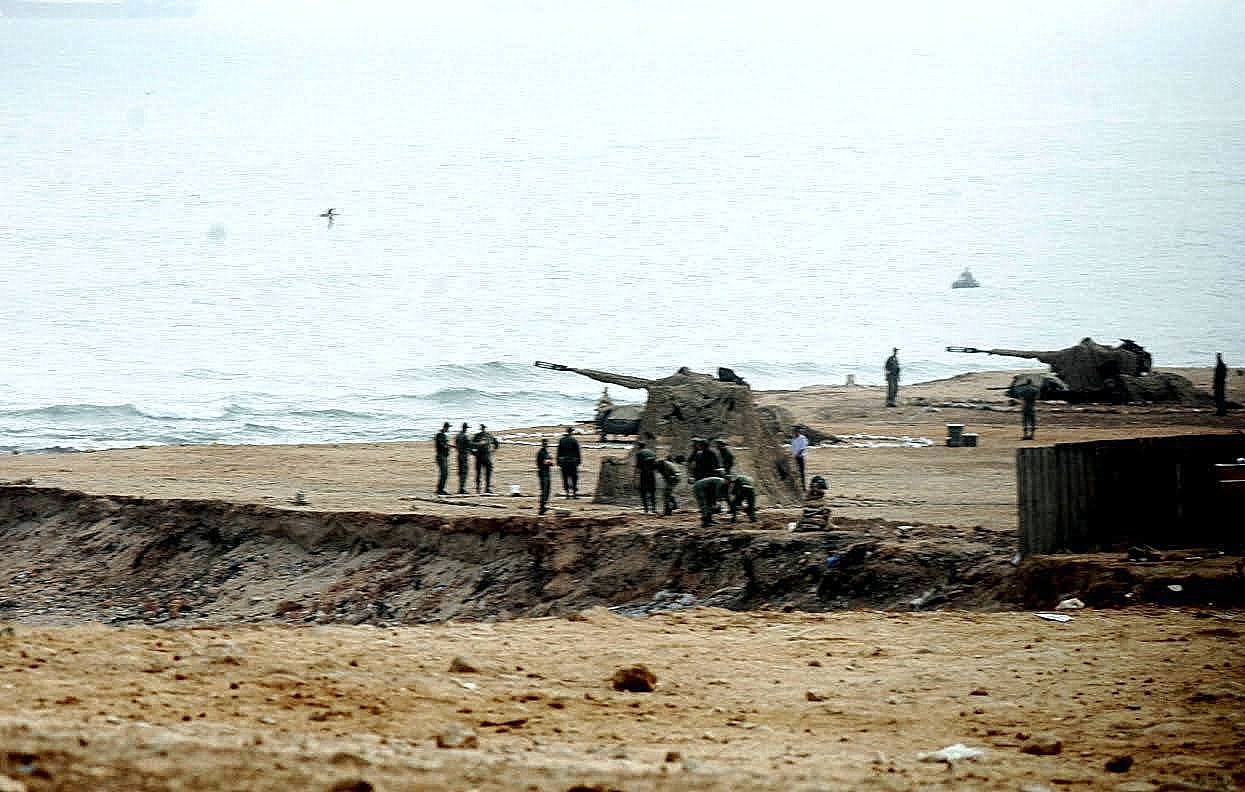 L'image montre une scène côtière où des militaires sont visibles sur une plage. On aperçoit des canons posés sur le sable, ainsi qu'un groupe de soldats qui semblent s'affairer autour. Le paysage est brumeux, avec une mer calme au fond et un ciel nuageux. L'atmosphère paraît sérieuse et militaire, avec une concentration sur les préparatifs ou les opérations en cours.