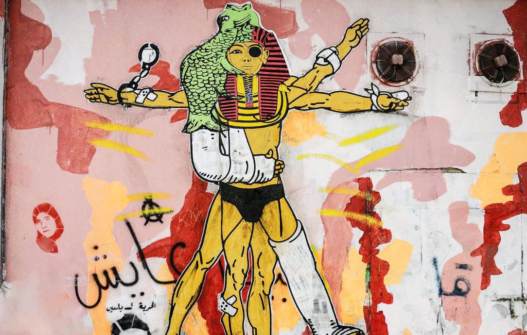 L'image représente une fresque murale colorée, incluant des éléments graphiques variés. Au centre, une figure humanoïde est stylisée avec des références à la culture égyptienne, incorporant des motifs inspirés des pharaons. Cette figure a plusieurs bras, symbolisant peut-être la force ou la multiplicités des rôles. Le fond est composé de formes abstraites et de couleurs vives, comme le rouge et le jaune, créant un contraste avec le personnage principal. On peut également apercevoir des inscriptions en arabe. L'ensemble véhicule une esthétique urbaine et contemporaine.