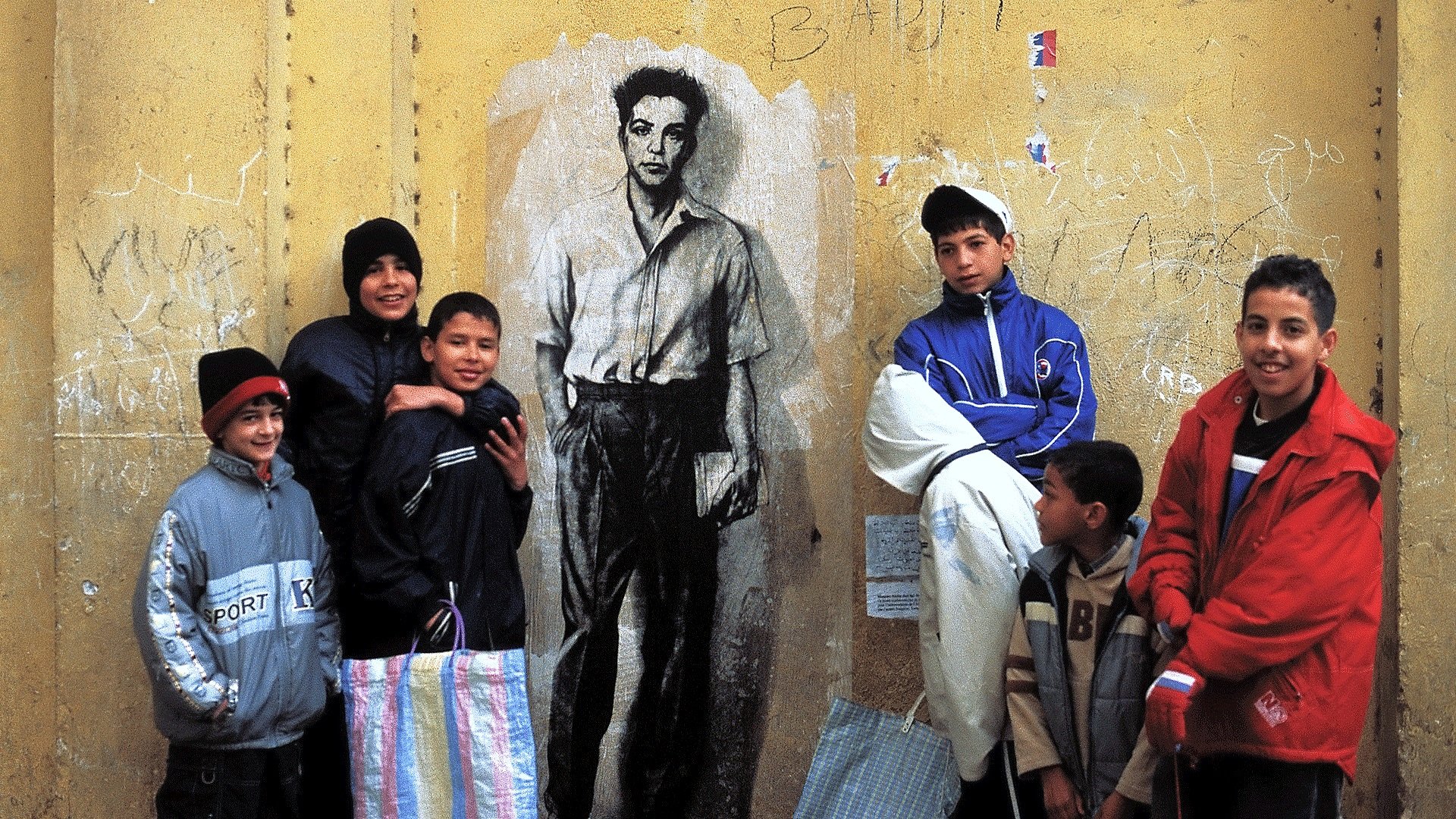 L'image montre un groupe de jeunes garçons rassemblés devant un mur de couleur jaune. Sur ce mur se trouve un dessin noir et blanc représentant un homme faisant face à la caméra. Les garçons, qui portent des vêtements variés et colorés, affichent des expressions diverses, allant du sourire à la curiosité. L'ensemble de la scène évoque un moment de camaraderie et de jeu dans un environnement urbain.