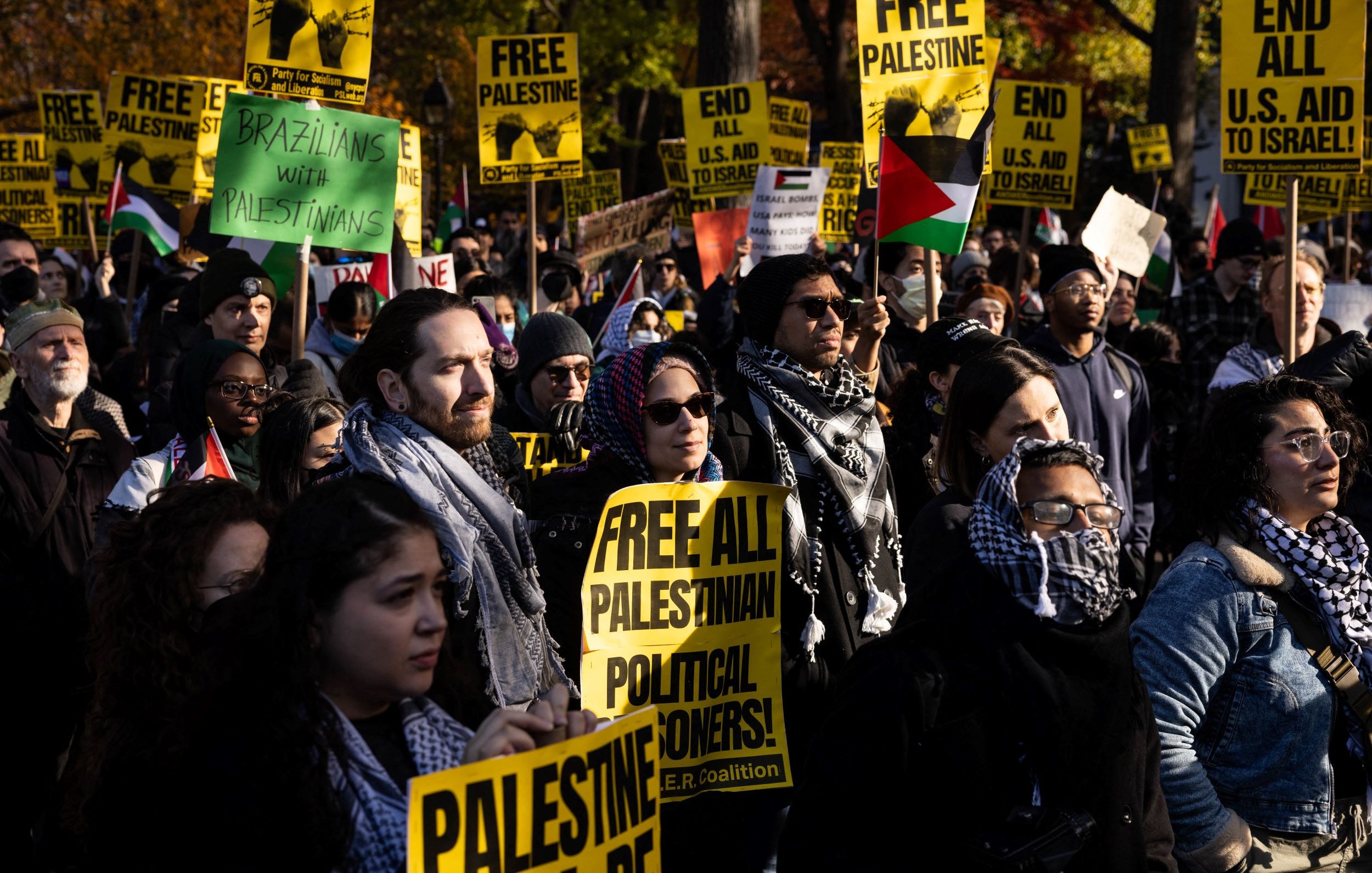 L'image montre une manifestation avec un grand nombre de personnes tenant des pancartes. Les pancartes portent des messages tels que "FREE PALESTINE" et "FREE ALL PALESTINIAN POLITICAL PRISONERS". Les participants sont vêtus de manière variée, certains portent des écharpes ou des keffiehs. On peut voir des drapeaux palestiniens en arrière-plan. L'ambiance semble être énergique et engagée, reflétant un fort sentiment de solidarité et de revendication pour les droits des Palestiniens.