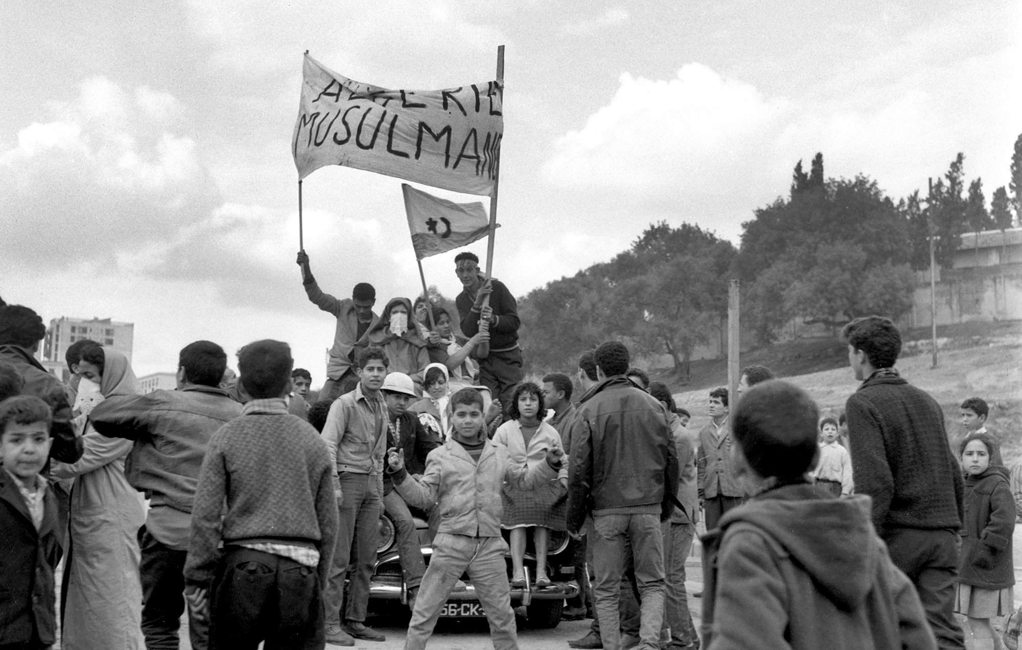L'image montre une scène de rue animée, probablement lors d'une manifestation ou d'un rassemblement. Des groupes de personnes, en majorité des jeunes, se rassemblent autour d'une voiture. Certains tiennent des drapeaux ou des banderoles sur lesquelles des messages sont affichés, exprimant des revendications. L'atmosphère semble dynamique et chargée d'émotion, avec des visages montrant diverses réactions. En arrière-plan, on aperçoit un paysage urbain avec des arbres et des bâtiments. Le noir et blanc de la photo accentue le caractère historique et sérieux de la scène.