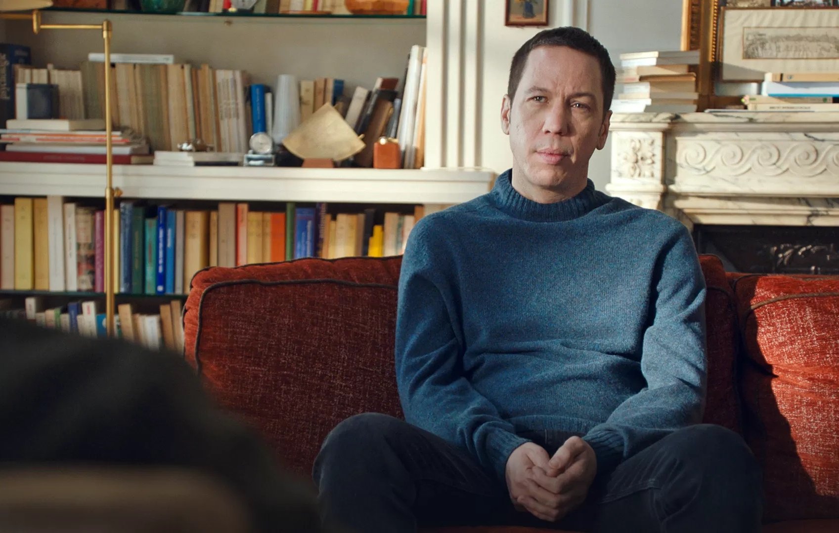 L'image montre un homme assis sur un canapé orange. Il porte un pull bleu et a une expression réfléchie. En arrière-plan, on peut voir une bibliothèque remplie de livres et quelques objets décoratifs. La lumière naturelle entre par une fenêtre, créant une ambiance chaleureuse et confortable dans la pièce. L'homme semble être en train d'écouter ou de participer à une conversation.