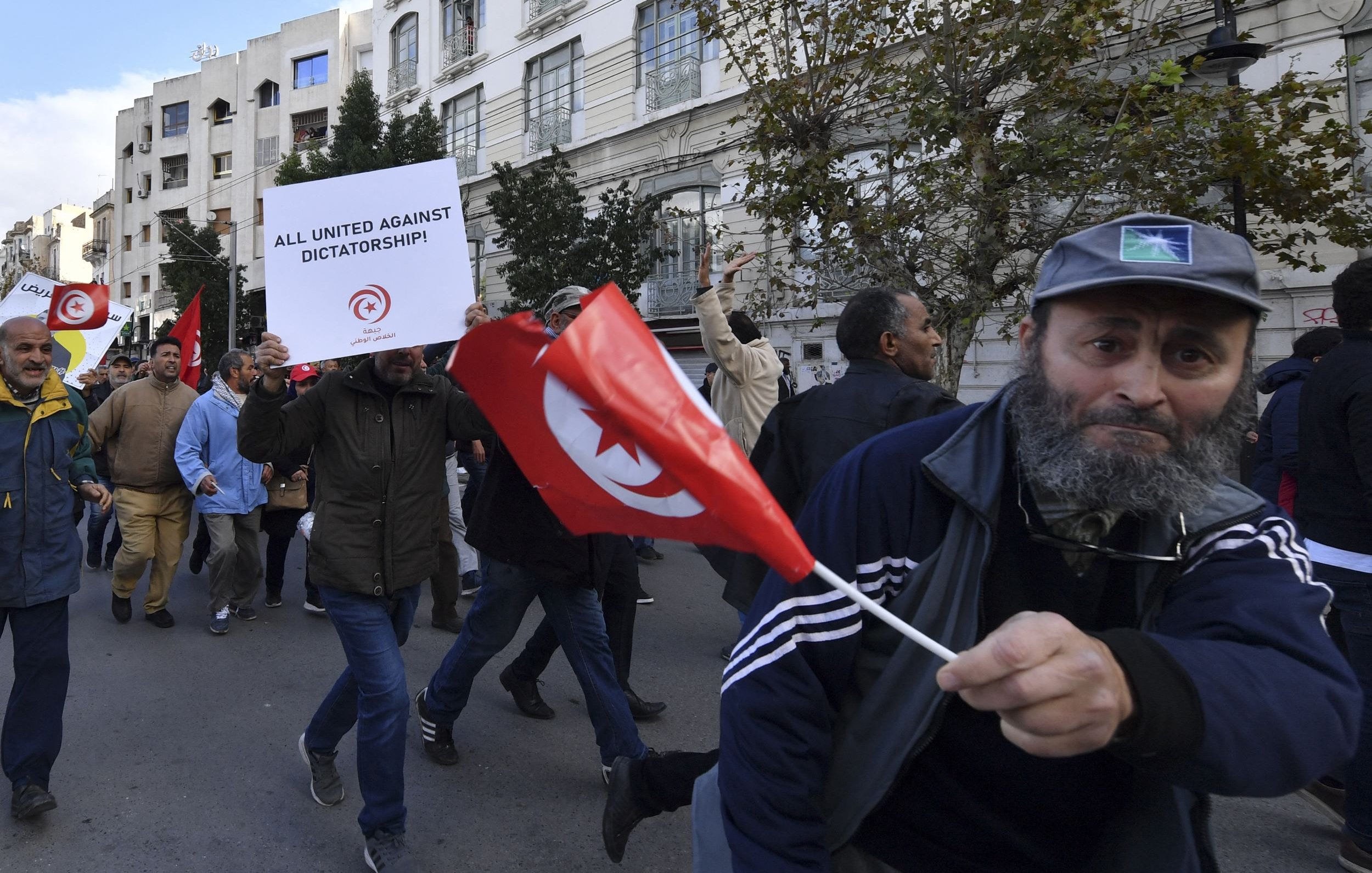 L'image montre une scène de manifestation avec des gens défilant dans la rue. Plusieurs manifestants portent des drapeaux tunisiens et tiennent des panneaux, dont l'un exprime un message contre la dictature. L'atmosphère semble énergique et engagée, avec des participants qui expriment leurs opinions et cherchent à faire entendre leur voix. Les bâtiments en arrière-plan reflètent un contexte urbain.