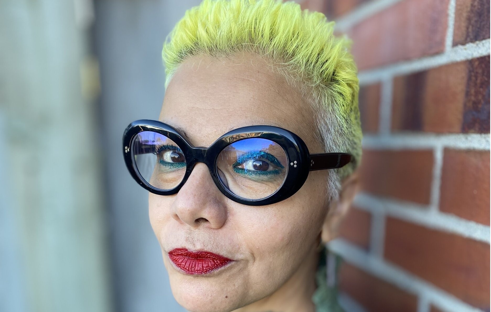 L'image montre une personne avec des cheveux très courts teints en jaune vif. Elle porte de grandes lunettes noires au style distinctif. Son maquillage comprend des lèvres rouge vif. Le fond présente un mur en briques, ce qui ajoute un contraste avec son look. Elle a une expression confiante et souriante.