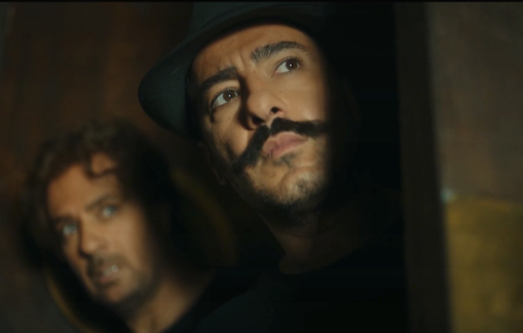 L'image montre deux hommes. L'un d'eux a une moustache prononcée et porte un chapeau, tandis que l'autre a les cheveux bouclés et semble inquiet. Ils regardent tous les deux vers la droite, comme s'ils étaient en train d'espionner ou de guetter quelque chose. L'éclairage est sombre, ce qui ajoute une atmosphère tendue et mystérieuse à la scène.