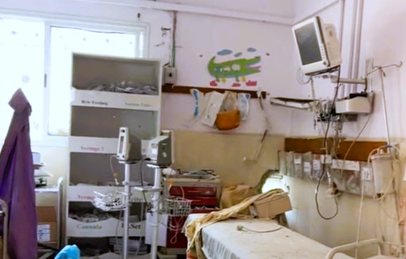 L'image montre une chambre d'hôpital qui a l'air un peu désordonnée et ancienne. On y voit un lit médical avec des draps, une étagère avec des boîtes, ainsi que du matériel médical. Un petit téléviseur est fixé au mur, et des illustrations colorées sont visibles sur les murs, ce qui ajoute une touche enfantine à l'environnement. La pièce semble nécessiter un peu de nettoyage et de modernisation.