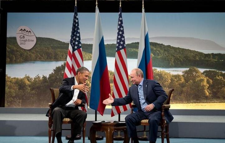 L'image montre deux dirigeants assis face à face dans un cadre formel. D'un côté, un homme souriant, vêtu d'un costume sombre avec une chemise claire, semble tendre la main vers son interlocuteur. De l'autre, un homme aux cheveux clairs, également en costume, répond avec un sourire et une expression chaleureuse. Derrière eux, on peut voir des drapeaux américains et russes, et un paysage naturel en arrière-plan. L'ensemble évoque une rencontre diplomatique ou un sommet entre ces deux pays.