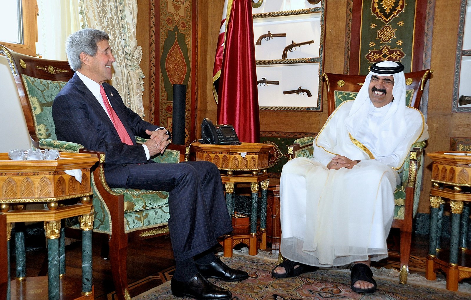 L'image montre deux hommes assis dans un bureau élégant, décoré avec des motifs riches et des couleurs vives. L'un des hommes porte un costume formel et sourit, tandis que l'autre est vêtu d'une tenue traditionnelle blanche, avec un chapeau noir. Ils semblent être en train de discuter dans une atmosphère amicale. En arrière-plan, on peut voir des objets décoratifs et des armes anciennes accrochées au mur, ajoutant à l'ambiance opulente de la pièce.