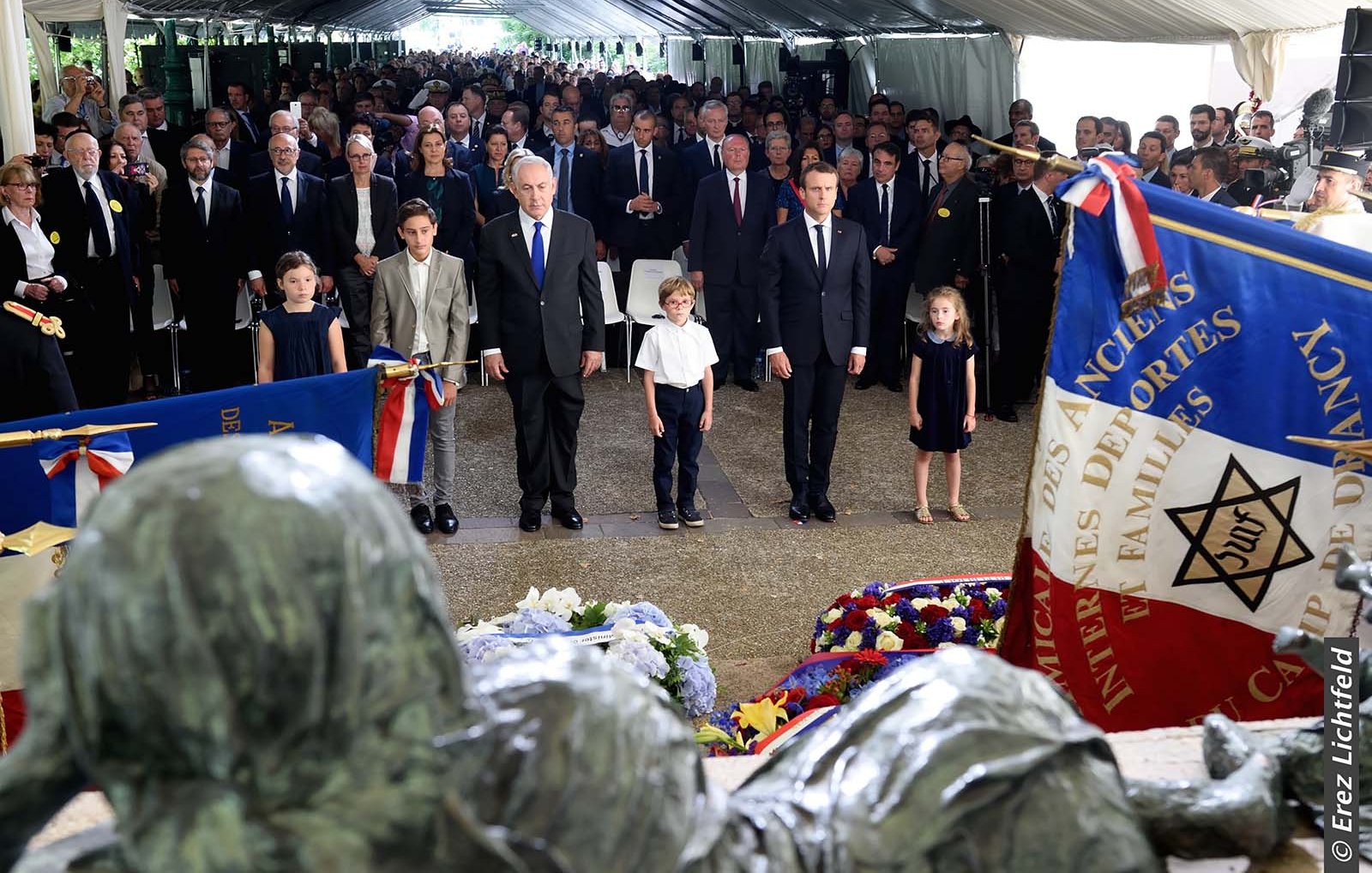 L'image montre une cérémonie commémorative avec un grand nombre de personnes rassemblées sous une tente. On peut voir des adultes en costumes et des enfants bien habillés. Au centre, deux hommes, dont l'un semble être un dirigeant, sont debout, les mains sur le cœur, en signe de respect. Il y a également des drapeaux et des gerbes de fleurs autour d'eux. L'ambiance semble solennelle et respectueuse, avec une attention particulière portée aux éléments de mémoire et d'honneur.