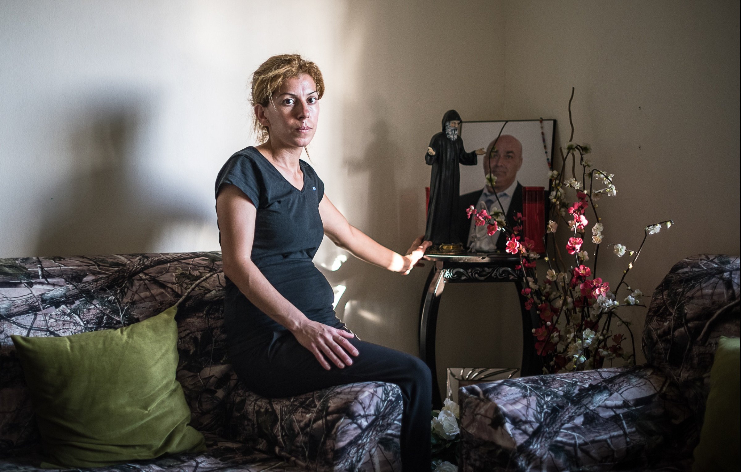 L'image montre une femme assise sur un canapé, portant une tenue noire. Elle est positionnée dans une pièce au mur neutre, avec un éclairage doux. À côté d'elle, il y a un petit meuble sur lequel se trouvent des objets décoratifs, ainsi qu'une photo encadrée d'un homme. Le décor du canapé est à motifs camouflage, et quelques fleurs artificielles ajoutent une touche de couleur à l'environnement. L'expression de la femme semble sérieuse, reflétant peut-être une émotion ou une réflexion profonde.
