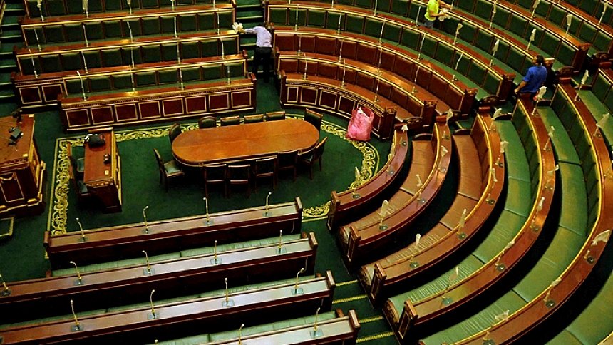 L'image montre l'intérieur d'une assemblée, probablement un parlement ou une salle de réunion officielle. On peut voir des rangées de sièges en bois oblongs disposés en arc de cercle, avec un grand bureau central ovale. Les bancs sont vides, ce qui donne une impression de calme. La décoration est sobre, avec des éléments en bois et un tapis vert. Deux personnes sont visibles, probablement en train de ranger ou d'entretenir l'espace. L'ambiance générale semble formelle et institutionnelle.