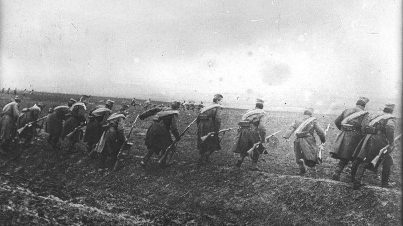 L'image montre un groupe de soldats marchant en formation sur un terrain accidenté. Ils portent des uniformes militaires, avec des chapeaux distinctifs, et semblent se déplacer dans une direction déterminée. Le paysage est marqué par un ciel nuageux et une ambiance sombre, suggérant peut-être un contexte de guerre ou de mouvement stratégique dans un environnement rural. Le sol est boueux, ce qui rend leur avance difficile. L'image est en noir et blanc, renforçant le caractère historique et poignant de la scène.
