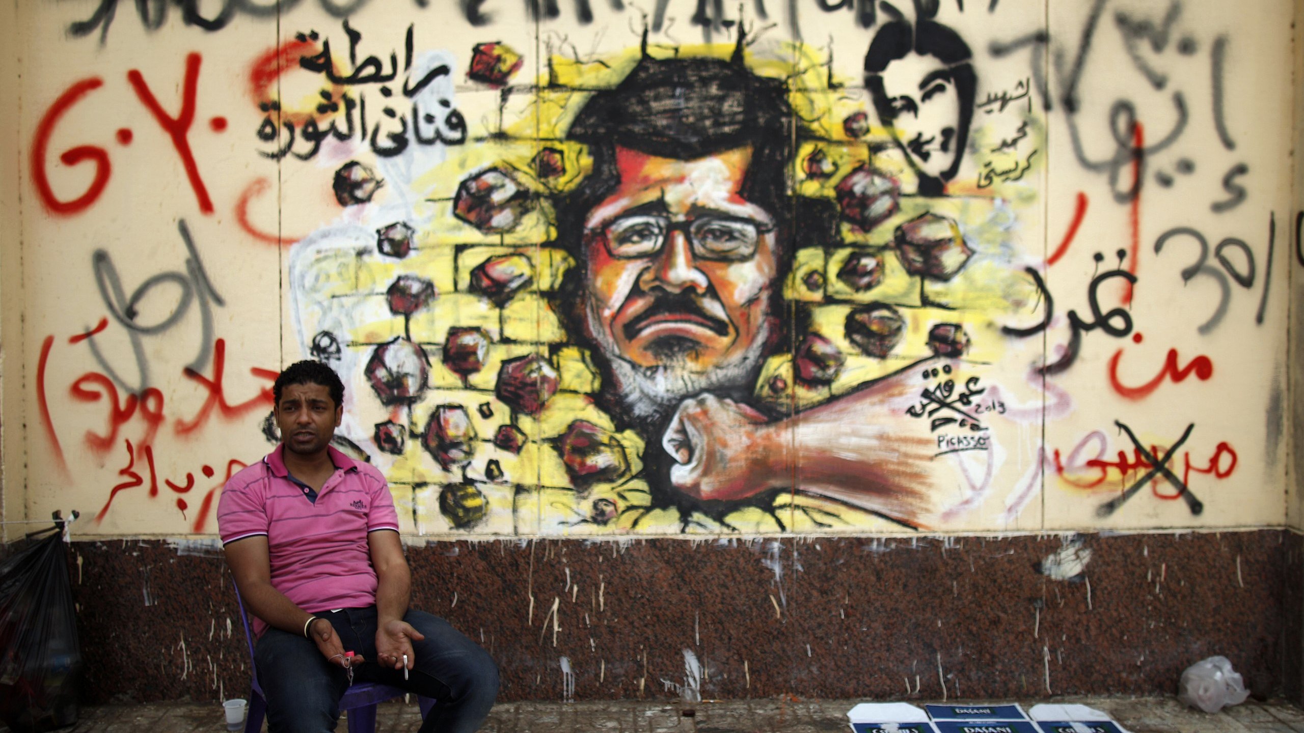 L'image montre un homme assis sur une petite chaise, devant un mur qui est recouvert d'une fresque murale. Cette fresco représente un portrait d'un homme au visage sérieux, avec des roches projetées autour de lui, symbolisant peut-être la résistance ou la lutte. Le mur est également couvert d'inscriptions en arabe et peut-être en d'autres langues, ajoutant un contexte politique ou social à la scène. L'environnement semble urbain, avec un sol en pavés et des déchets visibles à proximité.