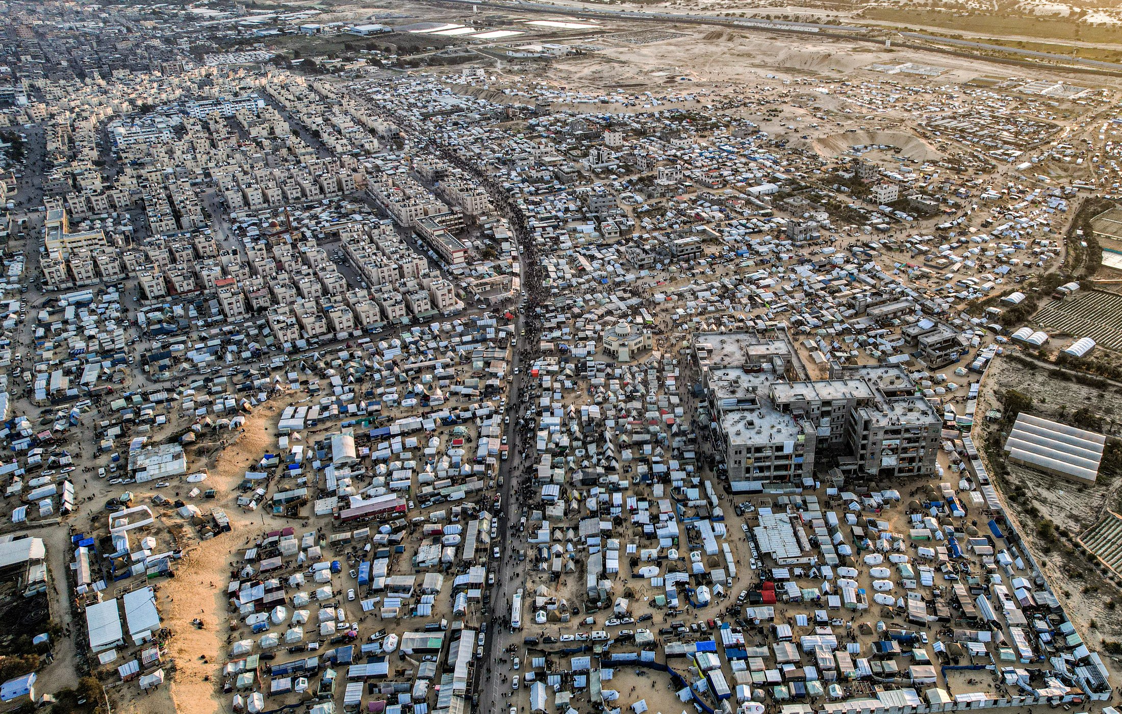 L'image montre une vue aérienne d'une vaste zone urbaine très densément peuplée, probablement une ville en développement ou un bidonville. On peut distinguer des habitations de différentes tailles et styles, de nombreuses structures temporaires, ainsi que des routes et des chemins qui traversent l'ensemble de la zone. La scène est marquée par un mélange de constructions durables et de tentes, suggérant une forte densité de population ainsi que des conditions de vie variées. En arrière-plan, on aperçoit des terrains plus dégagés, peut-être des zones agricoles ou industrielles.