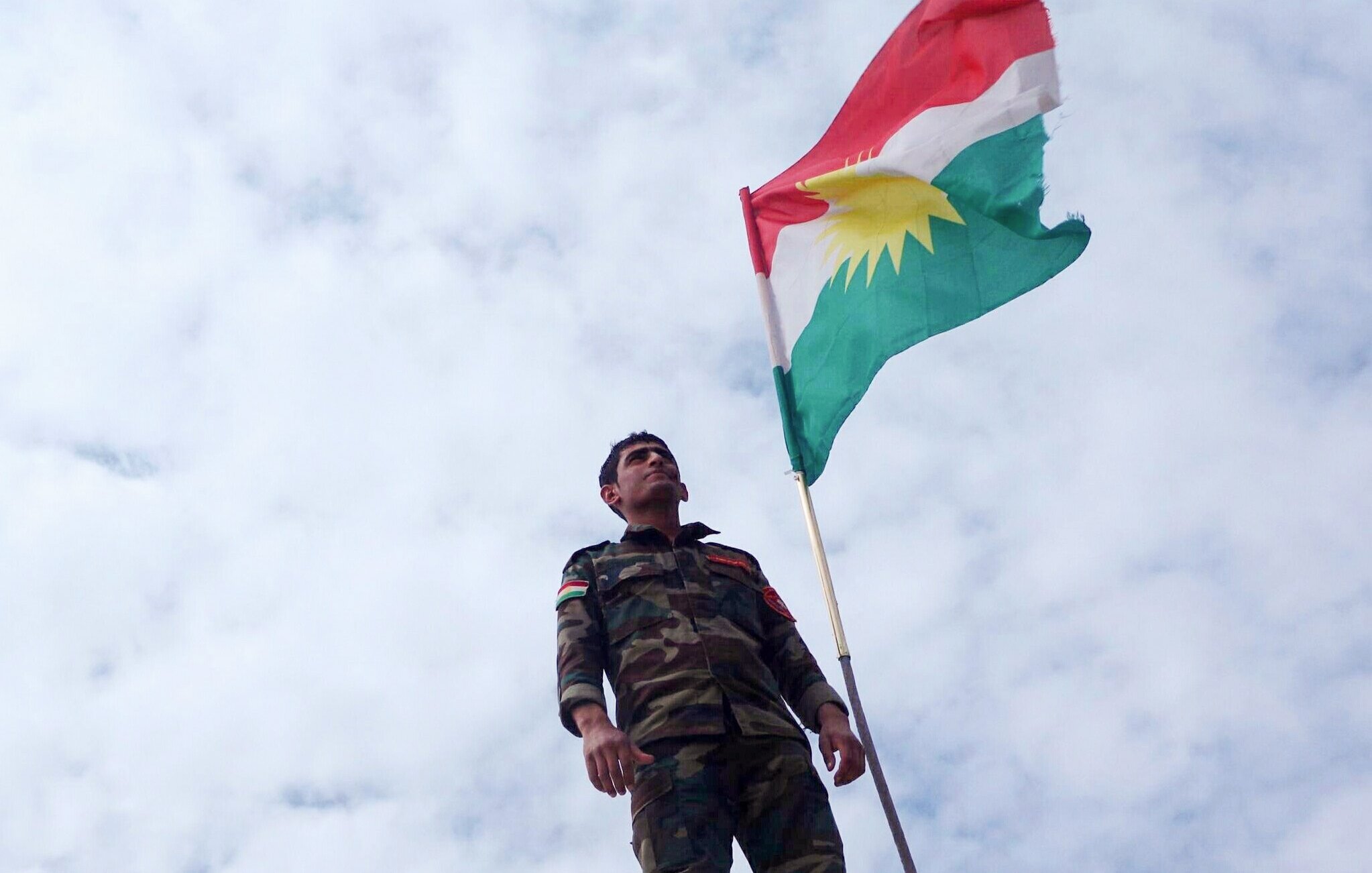 L'image montre un soldat portant un uniforme militaire, se tenant debout sous un drapeau kurde. Le drapeau présente les couleurs rouge, vert et blanc avec un soleil rayonnant au centre. Le ciel est nuageux, ajoutant une ambiance dramatique à la scène. Le soldat semble fier et déterminé, symbolisant l'engagement et l'identité kurde.