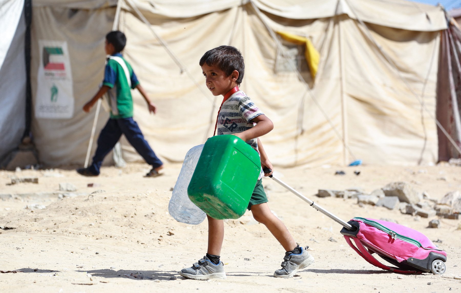 L'image montre un enfant marchant dans un environnement de camp, probablement un camp de réfugiés. Il tire une valise rose avec une poignée, tout en portant un récipient vert dans ses bras. En arrière-plan, on peut voir d'autres enfants et des tentes en toile, symbolisant un cadre de vie précaire. Le sol est poussiéreux et dénudé, ce qui reflète les conditions difficiles de l'endroit. L'enfant a un visage déterminé et semble concentré sur sa tâche.