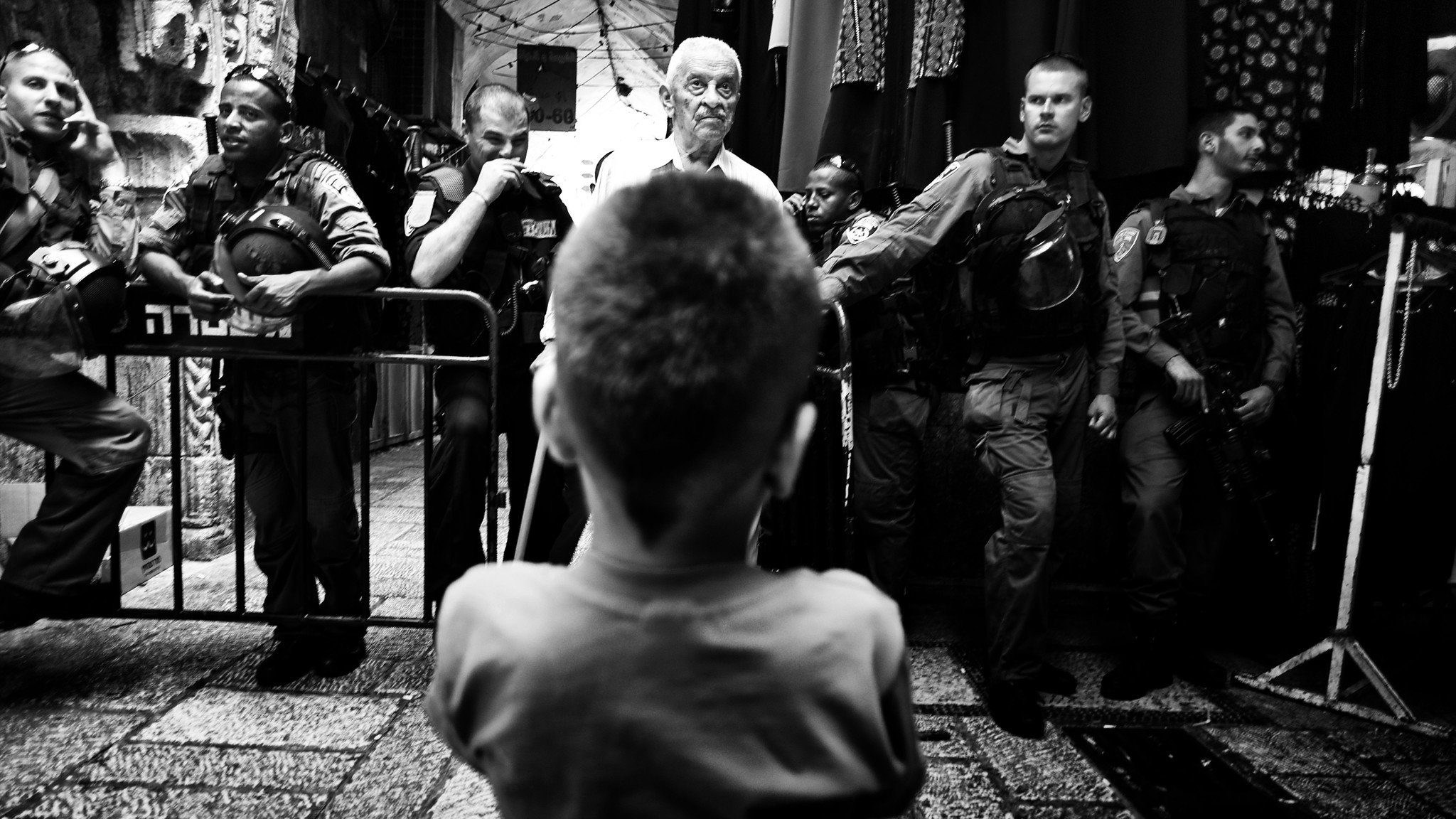 L'image montre un enfant vu de l'arrière, regardant vers un groupe de personnes, dont certaines semblent être des policiers ou des soldats. L'atmosphère est tendue, avec des barrières de sécurité en place et une composition en noir et blanc qui accentue le contraste entre les figures. L'enfant, central dans la scène, semble intrigué ou préoccupé par la situation qui se déroule devant lui. Les autres personnes sont alignées, probablement en position d'intervention.