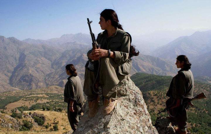 L'image montre trois personnes, probablement des combattants, dans un paysage montagneux. L'une d'elles est assise sur une pierre, tenant une arme, avec les jambes repliées. Les deux autres se tiennent debout en arrière-plan, également armées. Le décor est naturel, avec des montagnes majestueuses et une plaine en dessous, suggérant un environnement rural et sauvage. Les combattants portent des vêtements pratiques adaptés à leur environnement. L'ambiance de la scène semble sérieuse et déterminée.