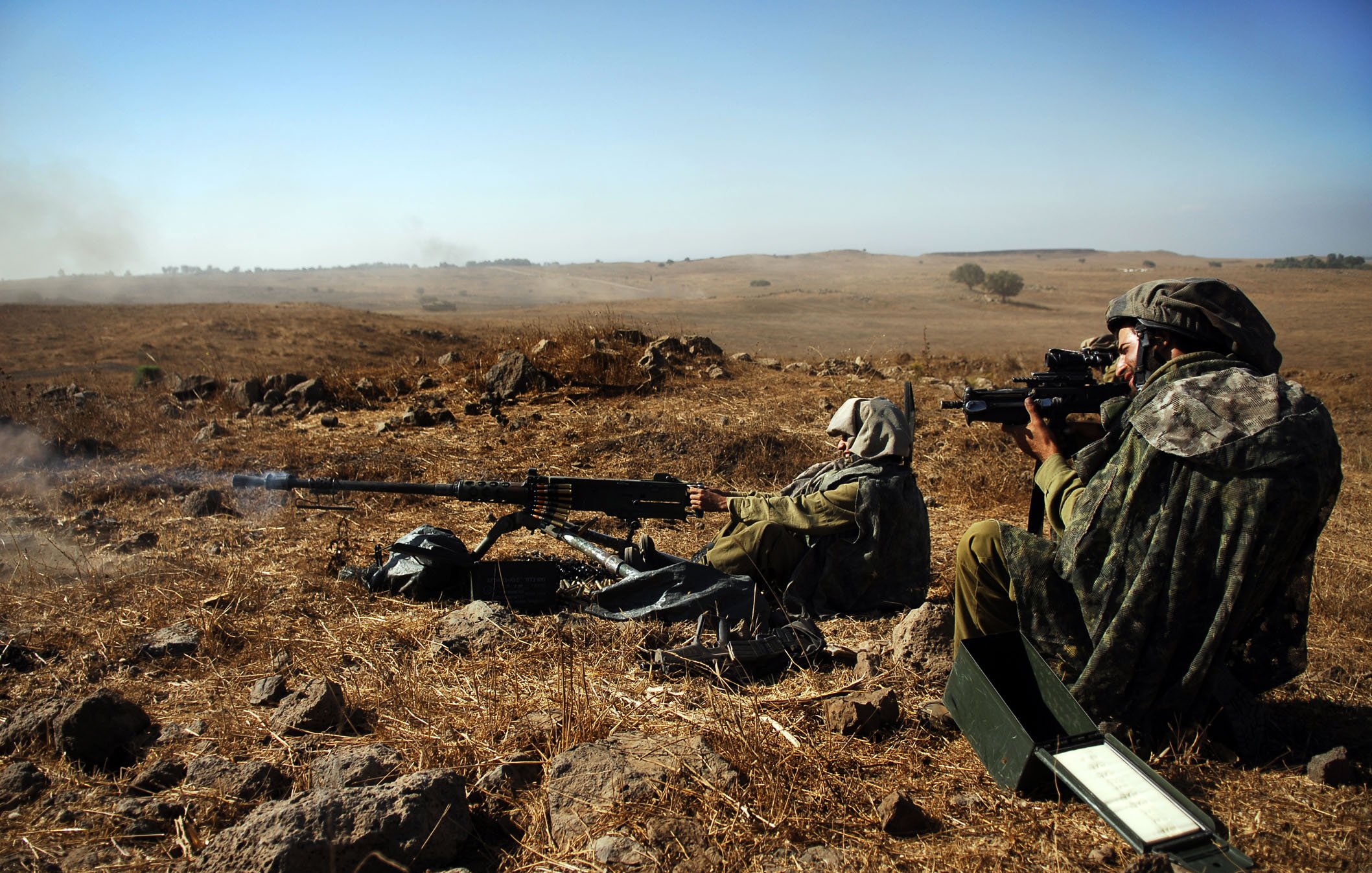 Sur l'image, on peut voir deux soldats en position de tir dans un paysage aride. Ils sont vêtus de tenue de camouflage et portent des casques. L'un des soldats est à genoux, prêt à tirer avec une mitrailleuse, tandis que l'autre est assis, observant à travers un dispositif de visée. Le terrain est composé de rochers et de terre sèche, et le ciel est dégagé, indiquant une journée ensoleillée. Cette scène évoque un environnement militaire austère et une préparation à l'action.