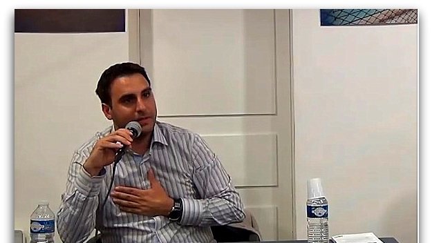 L'image montre un homme assis à une table, tenant un microphone. Il porte une chemise à rayures et semble s'exprimer avec conviction. Sur la table, il y a une bouteille d'eau et un gobelet, ainsi que des documents visibles. L'arrière-plan montre des affiches ou des œuvres, mais les détails ne sont pas clairement discernables.