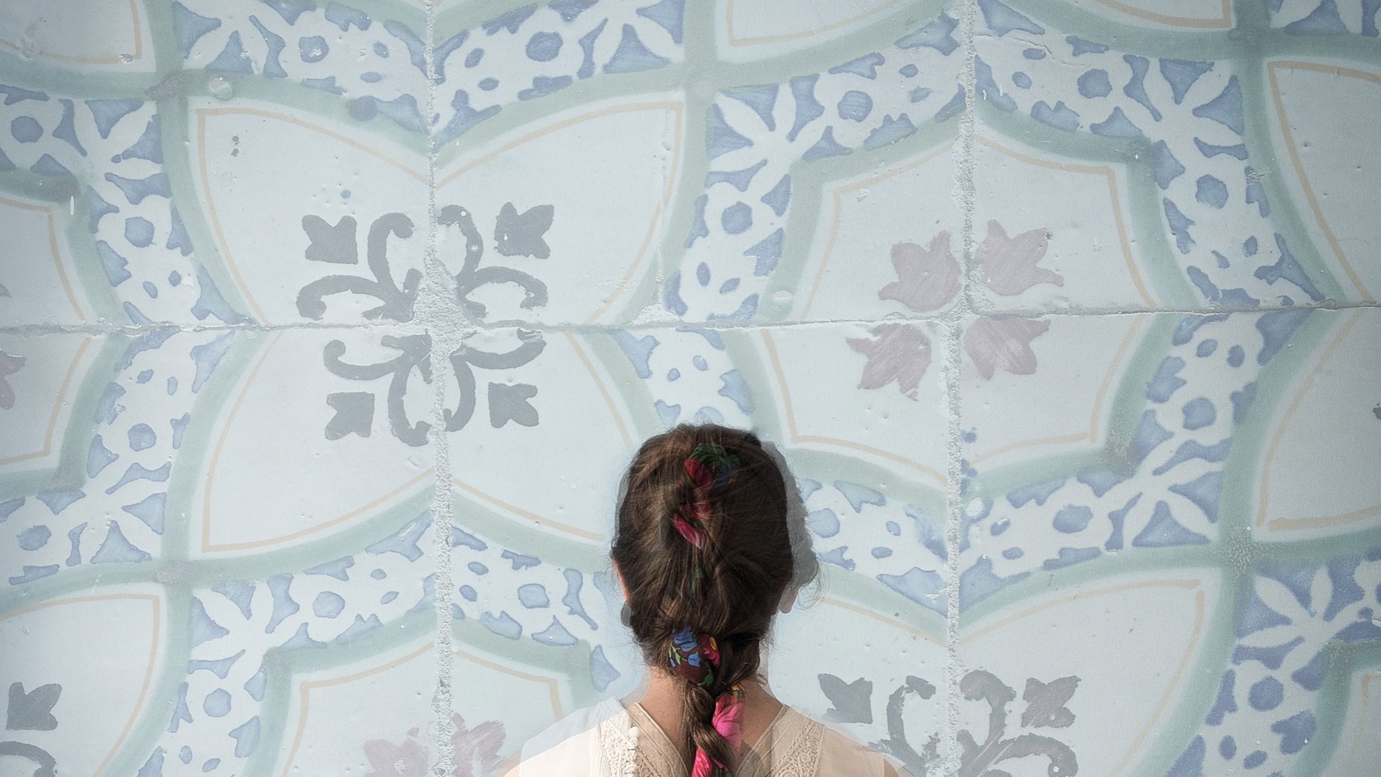 Cette image montre une personne de dos, portant un vêtement léger et délicat. Sa longue tresse, ornée de fleurs colorées, est visible et se détache sur un fond de motifs bleu clair. Les motifs sont ornés de formes géométriques et florales, créant une ambiance apaisante et artistique. L'ensemble évoque une atmosphère de calme et de beauté.