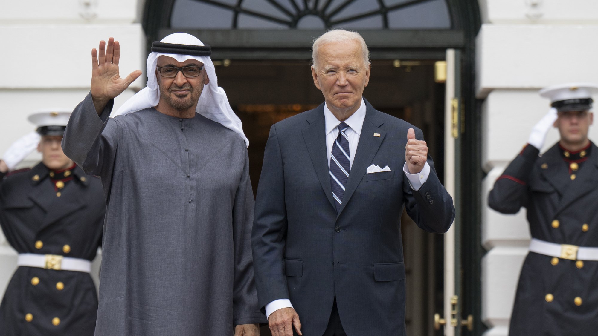 L'image montre deux hommes se tenant à l'extérieur d'un bâtiment, probablement la Maison Blanche. L'un des hommes est vêtu d'une robe traditionnelle, un keffieh sur la tête, et fait un geste de la main, tandis que l'autre porte un costume avec une cravate. Derrière eux, des membres des forces armées en uniforme saluent. L'ambiance semble formelle, suggérant un événement diplomatique ou une rencontre officielle.