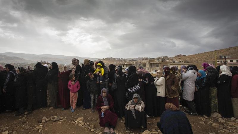 L'image montre un groupe de femmes rassemblées, probablement en attente de quelque chose. Elles portent des vêtements sombres, et certaines tiennent des enfants. Le ciel est nuageux, ce qui donne une ambiance sombre à la scène. En arrière-plan, on aperçoit des maisons sur une colline, soulignant un environnement rural ou de village. L'attente et la solidarité féminine semblent être des thèmes présents dans cette image.