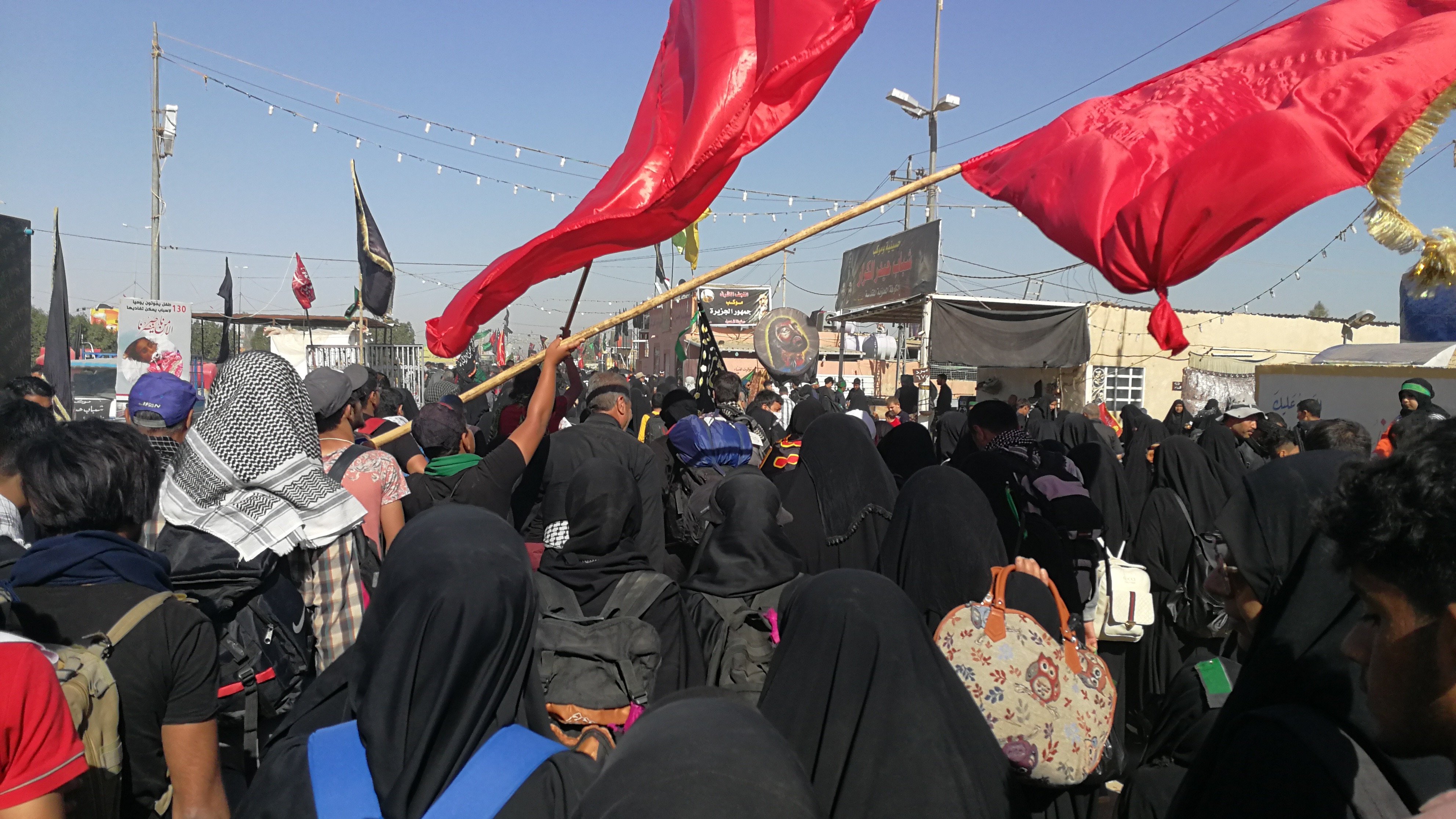 L'image montre une grande foule de personnes rassemblées, principalement vêtues de noir, ce qui suggère un événement commémoratif ou religieux. Plusieurs personnes tiennent des drapeaux rouges, et l'atmosphère semble être celle d'une manifestation ou d'un rassemblement significatif. Les participants portent des vêtements traditionnels et il y a des décors typiques de ce genre d'événement en arrière-plan, comme des bannières et éventuellement des symboles religieux. Le ciel est clair, ce qui indique une journée ensoleillée.