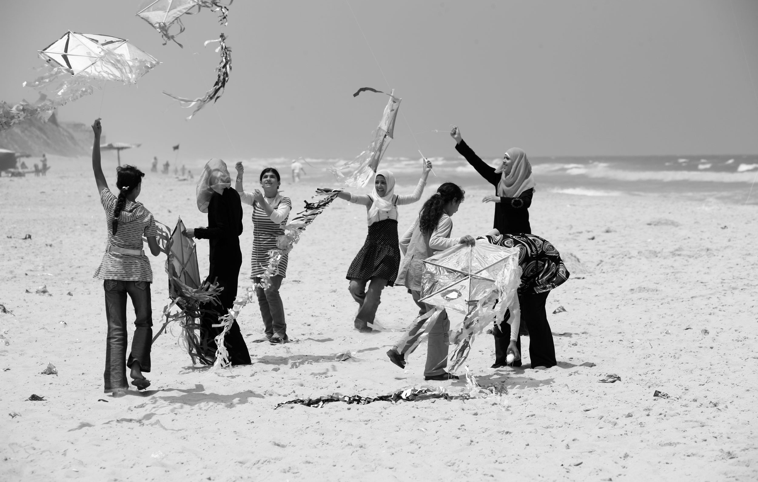 L'image montre un groupe de femmes sur une plage, s'amusant à faire voler des cerfs-volants. Certaines d'entre elles sont en train de tenir ou de manipuler des cerfs-volants, tandis que d'autres semblent rire et profiter du moment. L'environnement est ensoleillé, avec des vagues en arrière-plan. La scène dégage une atmosphère joyeuse et festive, mettant en avant des interactions sociales et des activités de plein air.