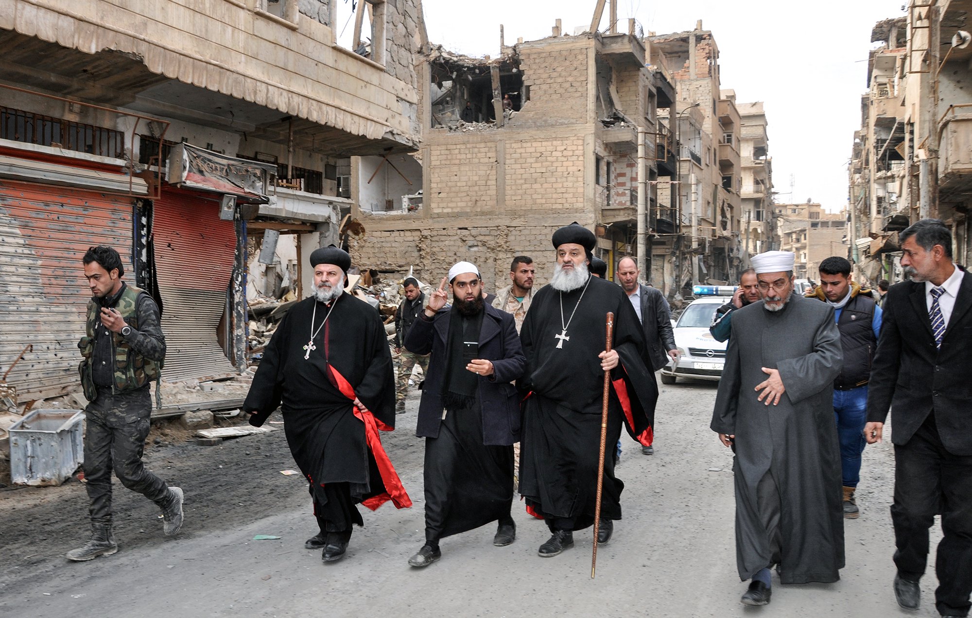 L'image montre un groupe de personnes marchant dans une rue endommagée. Il y a des bâtiments en ruines sur les côtés, indiquant une zone touchée par des conflits. Parmi le groupe, plusieurs hommes portent des vêtements traditionnels, notamment des robes noires et des croix, ce qui suggère qu'ils pourraient être des leaders religieux. Certains d'entre eux tiennent des bâtons. En arrière-plan, des militaires et des véhicules sont visibles, ajoutant à l'ambiance de la scène.