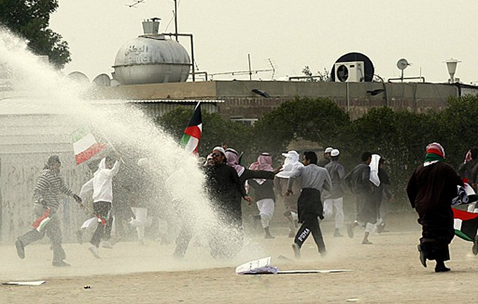 L'image semble montrer une scène de manifestation ou de rassemblement. On peut voir un groupe de personnes courant, certains tenant des drapeaux. Il y a également un jet d'eau qui pourrait provenir d'un canon à eau, suggérant une intervention des forces de l'ordre. L'arrière-plan comporte des bâtiments et un environnement rural, avec de la poussière soulevée par le mouvement. L'atmosphère semble tendue.