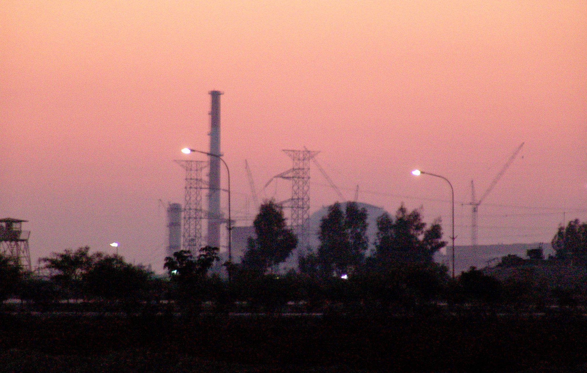 L'image montre une scène de crépuscule avec un ciel teinté de rose et d'orange. Au premier plan, on peut voir des arbres ainsi que des lampadaires allumés. En arrière-plan, se dressent des structures industrielles, notamment des cheminées et des pylônes électriques, ainsi que des grues de construction, ce qui suggère une activité industrielle en cours. L'atmosphère est calme et paisible, malgré la présence des installations.