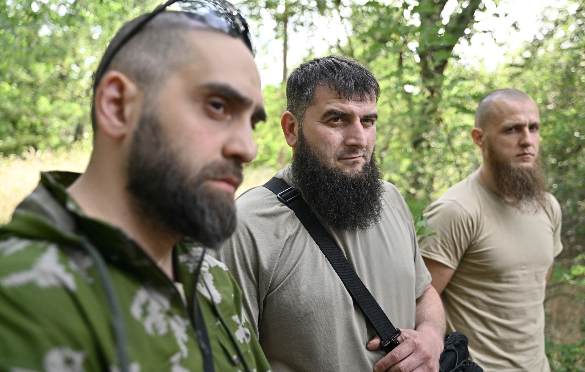 L'image montre trois hommes se tenant côte à côte dans une forêt. Tous trois portent des vêtements utilitaires, et chacun d'eux a une barbe. Leurs expressions sont sérieuses, et l'éclairage naturel met en valeur leur présence dans ce cadre boisé. On peut observer des arbres en arrière-plan, ce qui crée une ambiance naturelle.