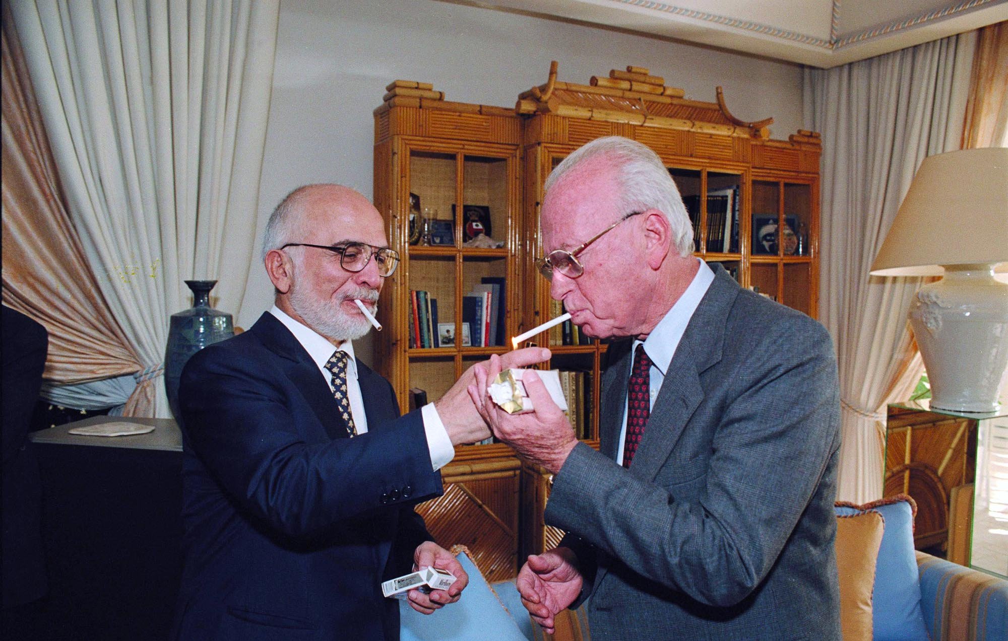 L'image montre deux hommes, chacun tenant une cigarette. L'un des hommes, vêtu d'un costume sombre et portant des lunettes, allume la cigarette de l'autre homme, qui porte également un costume et a une barbe grise. Ils se tiennent dans une pièce élégante, avec des meubles raffinés et des étagères en arrière-plan. L'atmosphère semble conviviale et intime.