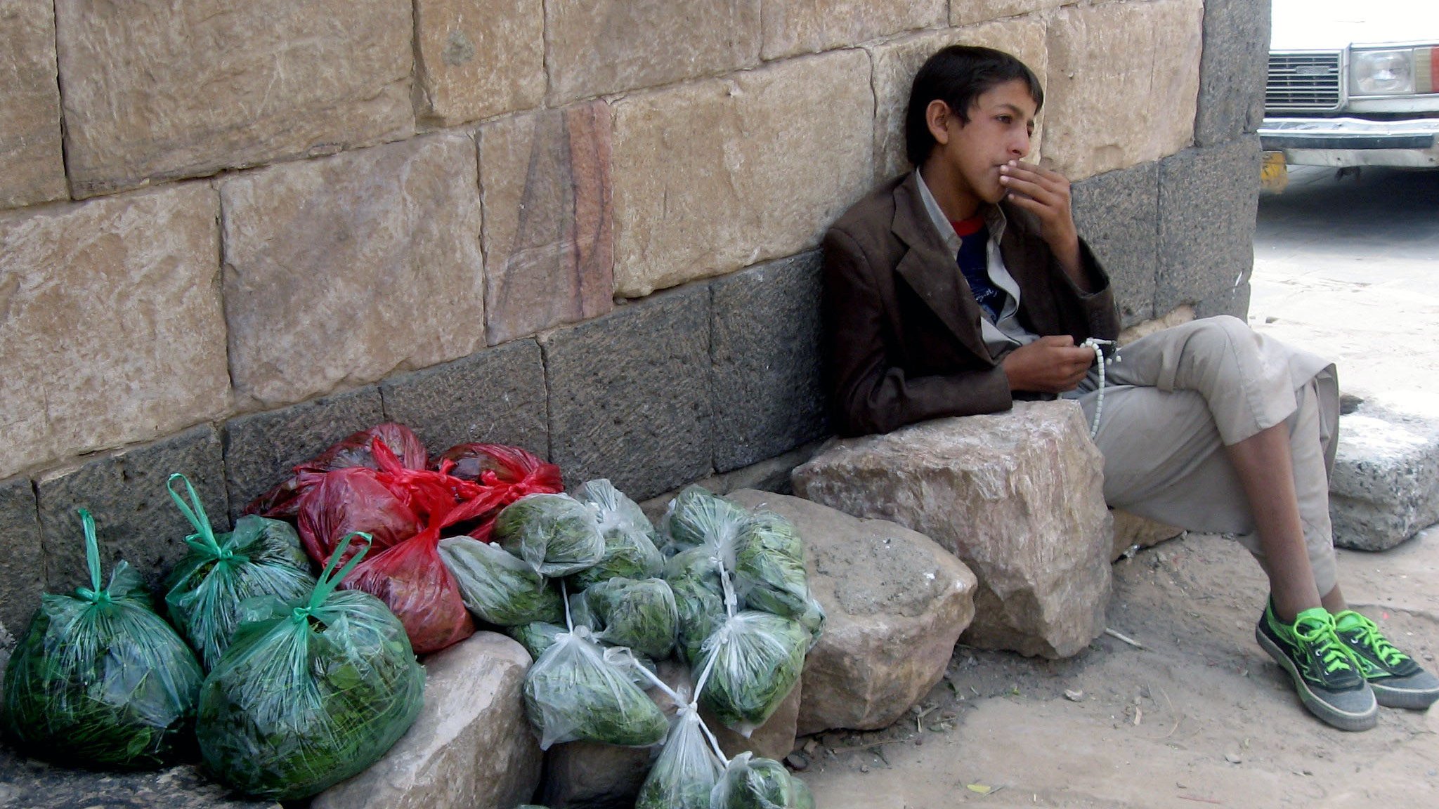 L'image montre un jeune garçon assis contre un mur en pierre. Il porte une veste et des pantalons clairs, et il semble fumer. À côté de lui, il y a plusieurs sacs transparentes contenant probablement des légumes ou des herbes, empilés sur le sol. Les sacs sont de différentes couleurs, principalement verts avec quelques rouges, et ils sont disposés sur des roches à ses côtés. L'environnement semble urbain, avec une route visible en arrière-plan.