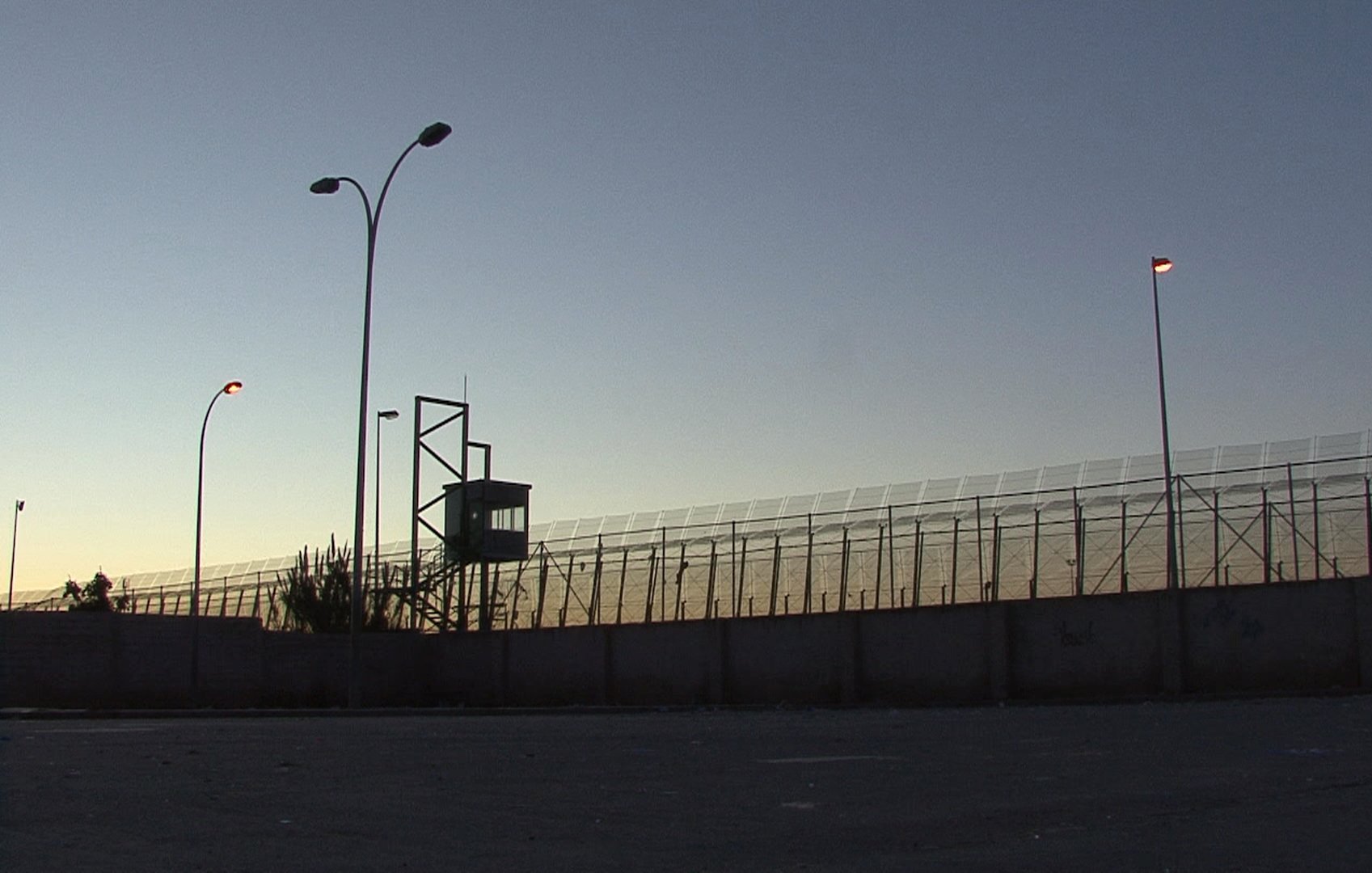 L'image montre une clôture de sécurité, probablement d'une prison ou d'une zone contrôlée. On aperçoit une tour d'observation au fond, illuminée par des lampadaires. La scène se déroule au crépuscule, avec un ciel qui passe des teintes orange et bleues, créant une atmosphère à la fois calme et tendue. Le mur en béton à la base de la clôture souligne le caractère restrictif de l'endroit.