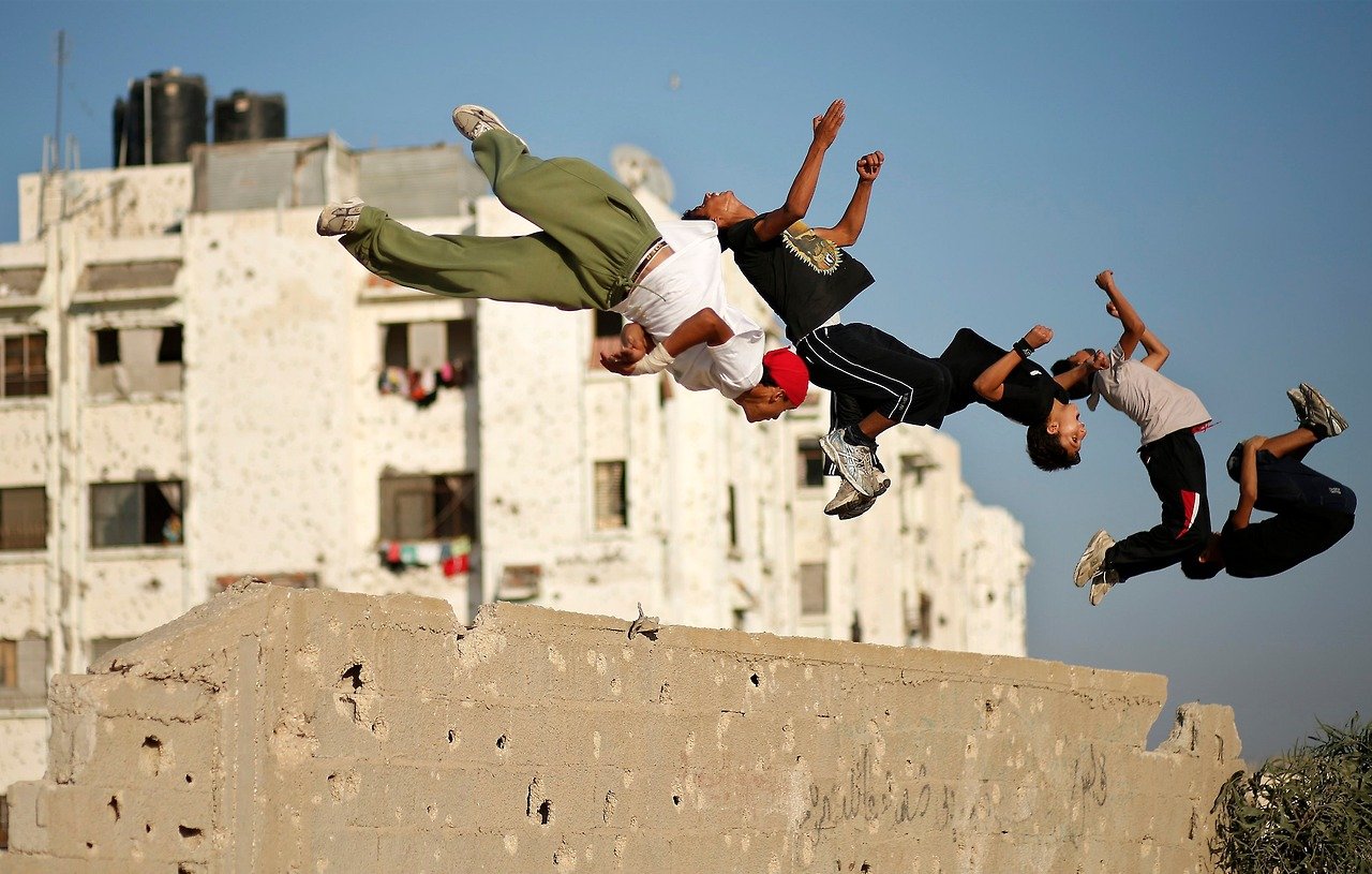 L'image montre un groupe de jeunes en train de sauter par-dessus un mur. Ils semblent s'amuser à réaliser des acrobaties, témoignant d'une grande énergie et d'une certaine audace. En arrière-plan, on aperçoit des bâtiments avec des murs marqués, ce qui donne une idée de l'environnement urbain. Le ciel est clair, ce qui contraste avec les structures des bâtiments. L'ambiance générale de l'image est dynamique et vivante, capturant un moment de loisirs et de camaraderie entre les adolescents.