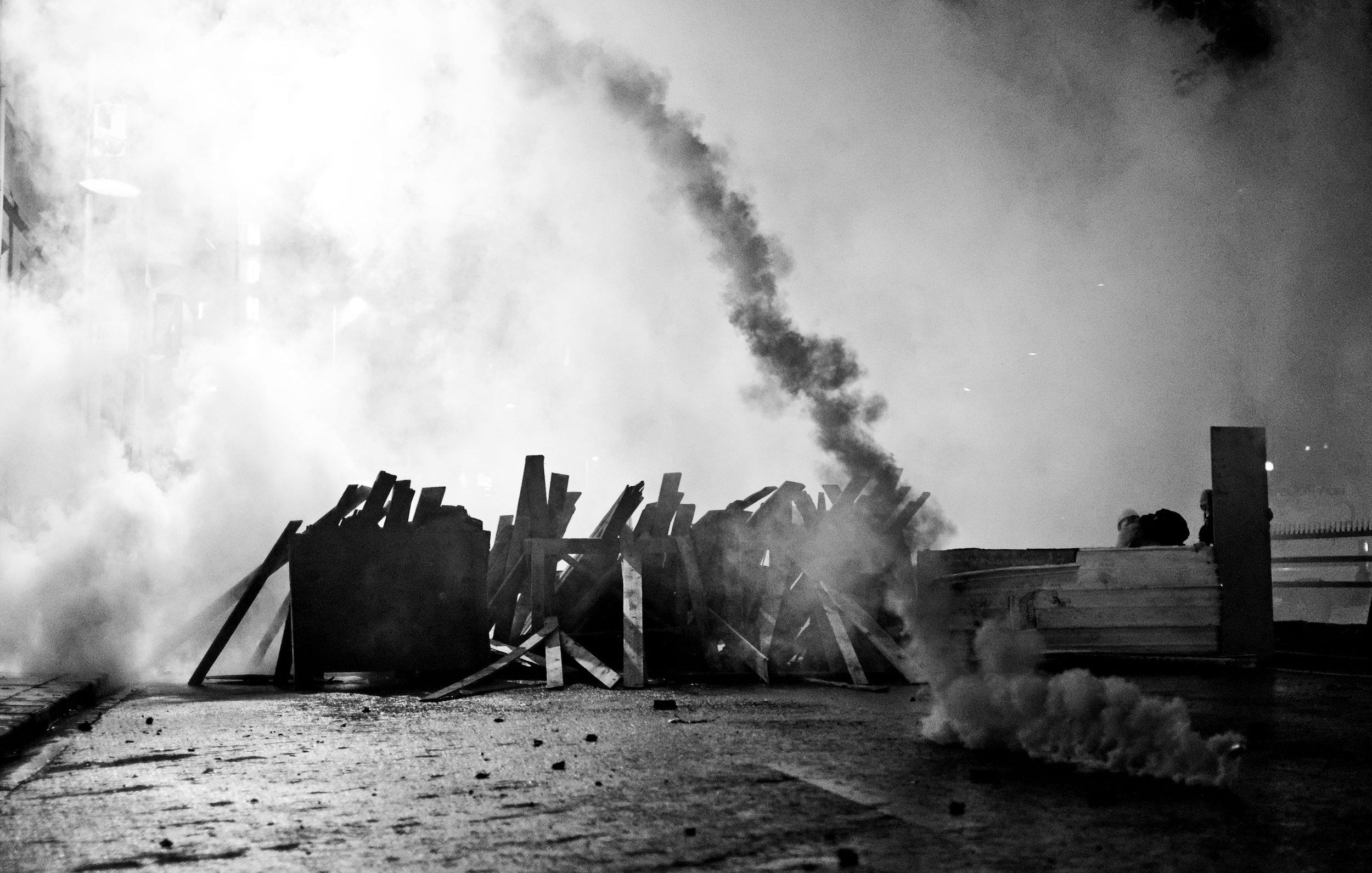 L'image montre une scène de tension, probablement associée à une manifestation ou à des troubles civils. On y voit des barricades faites de planches de bois, qui semblent être disposées sur une route. Une épaisse fumée s'élève dans l'air, créant une atmosphère sombre et chaotique. La monochromie de la photo ajoute à l'intensité de la scène, soulignant le contraste entre les ombres et la lumière. Des éléments comme des fumigènes ou des projectiles peuvent également être présents, suggérant une confrontation entre manifestants et forces de l'ordre. L'ensemble évoque une situation de conflit et de désobéissance civile.