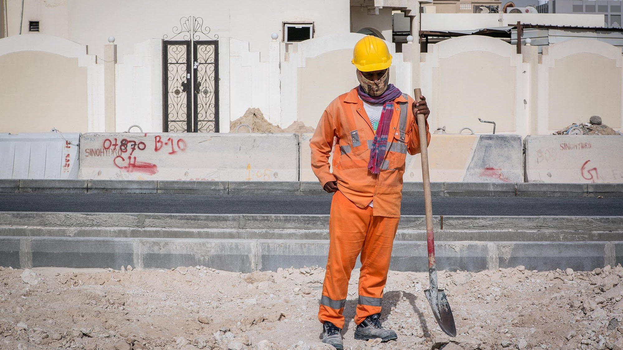 L'image montre un homme qui travaille sur un chantier de construction. Il porte un uniforme orange, ainsi qu'un casque de sécurité jaune. Il semble tenir une pelle, regardant vers le sol avec une posture réfléchie. En arrière-plan, on peut voir des murs de construction et des graffitis sur des surfaces environnantes. Le site semble en cours de développement, avec des matériaux de construction dispersés au sol.